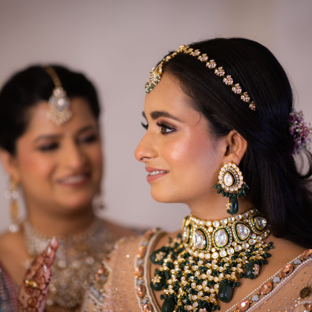 Two women dressed in traditional Indian wedding attire, adorned with elaborate jewelry and flowers, smiling at each other.