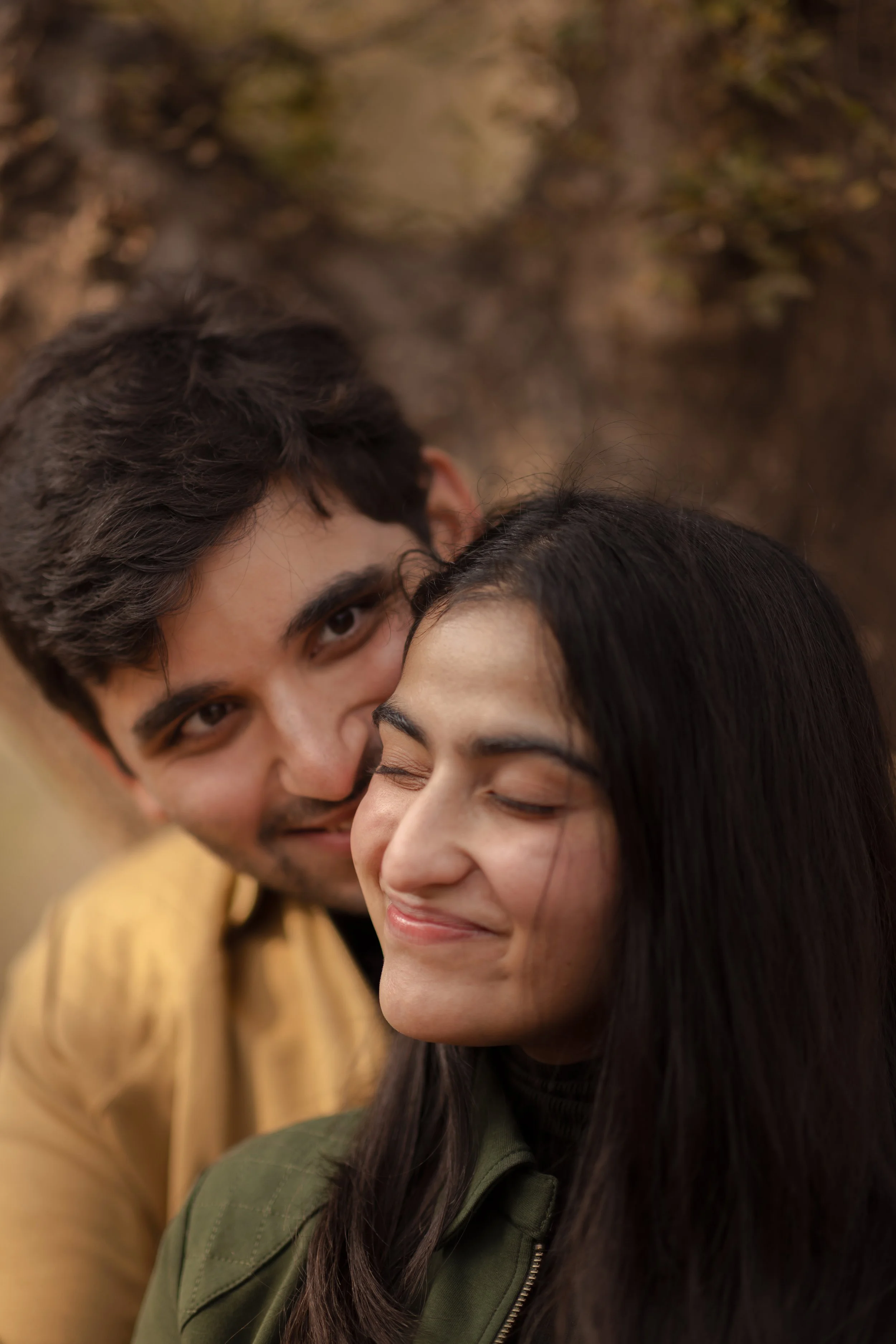 A young man and woman sharing a tender moment outdoors, with the man smiling and leaning close to the woman who has her eyes closed and a gentle smile.