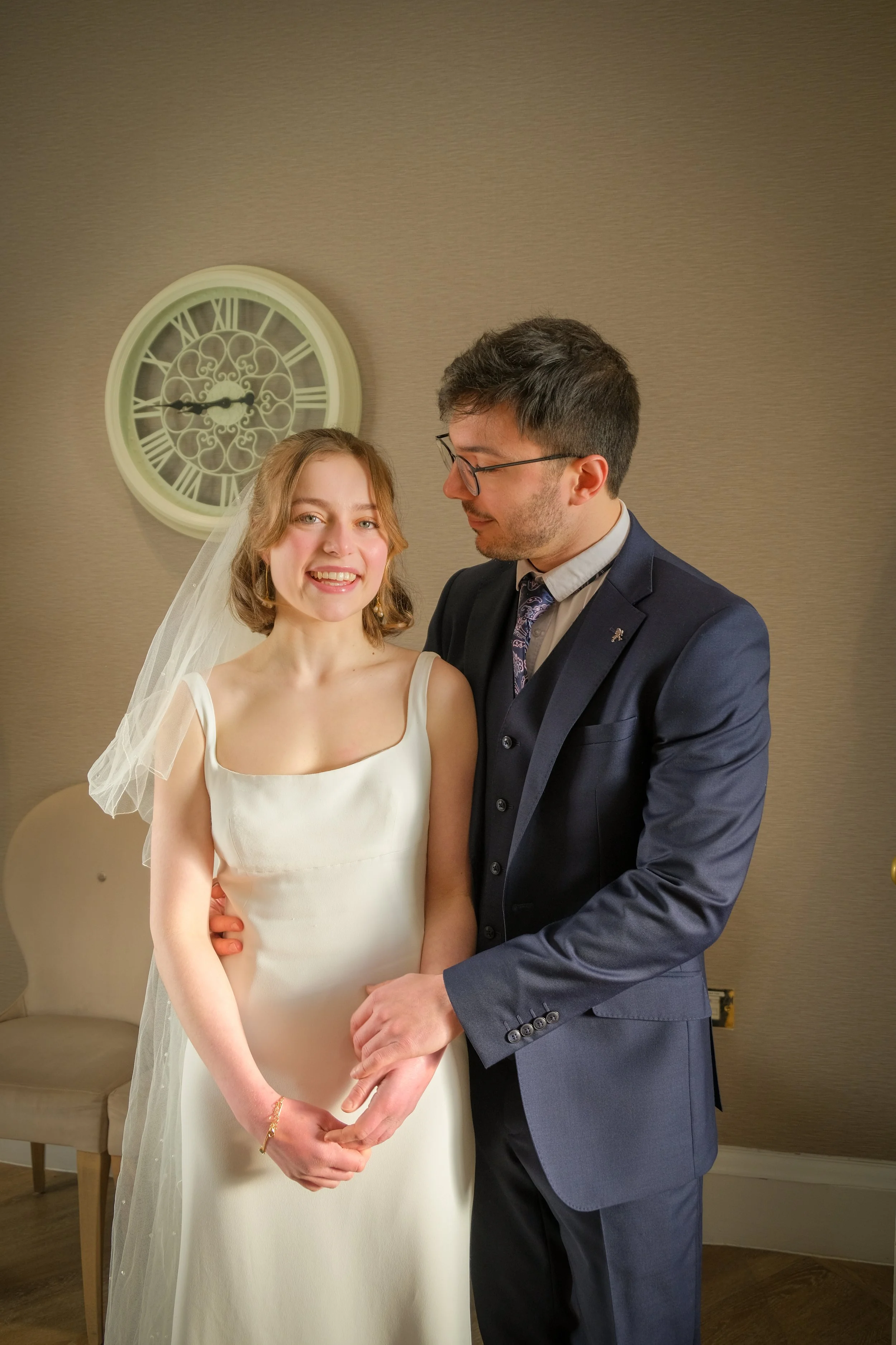 A woman in a white wedding dress and veil smiling and holding hands with a man in a dark blue suit, indoors with a beige wall, a clock, and a chair in the background.