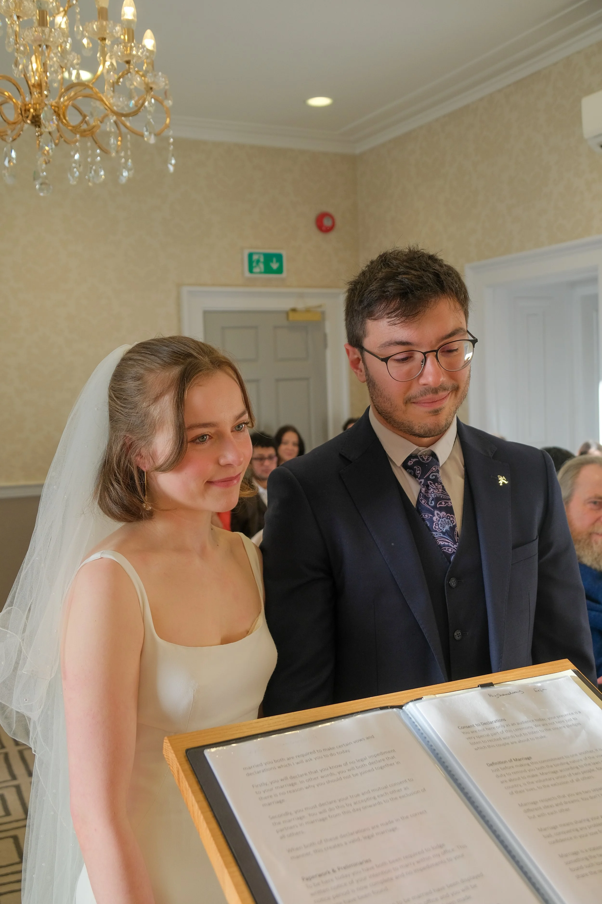 A bride and groom standing together during their wedding ceremony, looking at a document or script on a stand.