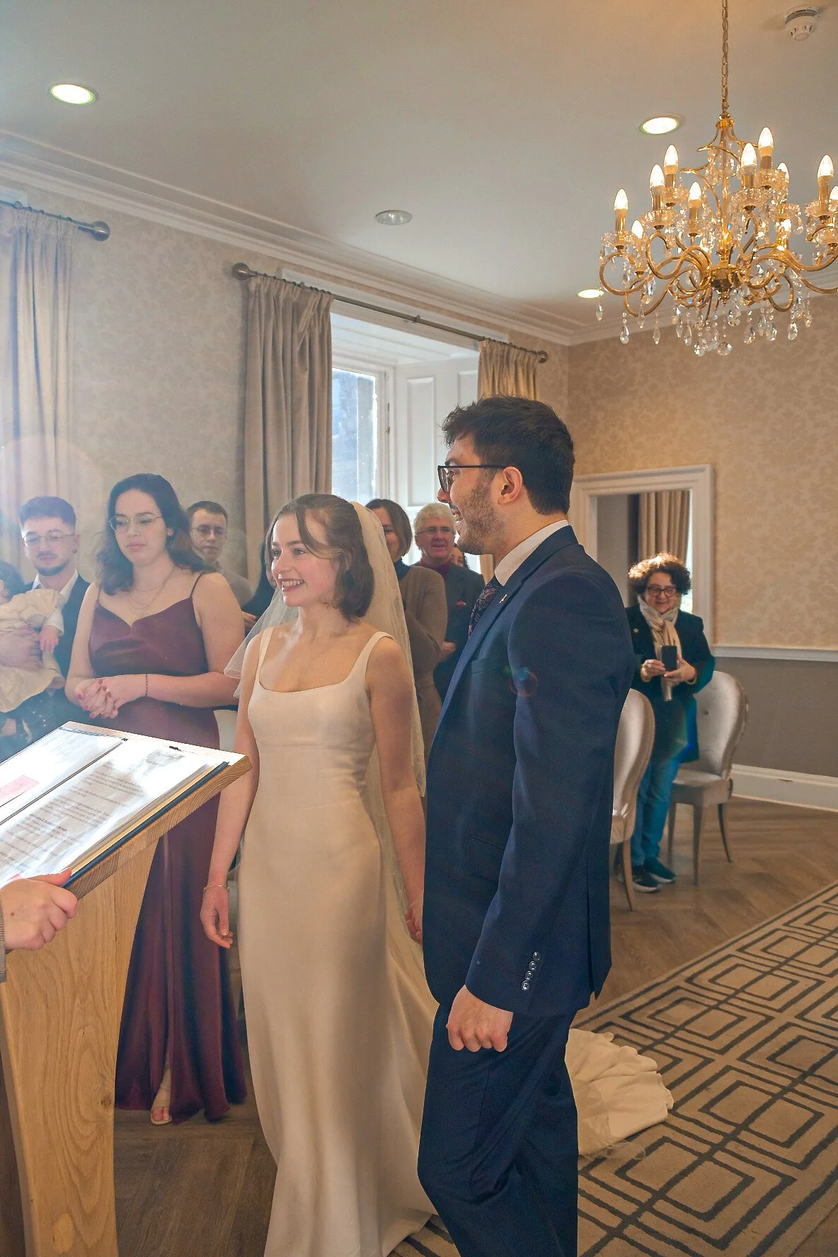 A wedding ceremony taking place indoors with the bride and groom standing in front of an officiant, surrounded by guests in a decorated room with curtains, a chandelier, and an ornate rug.