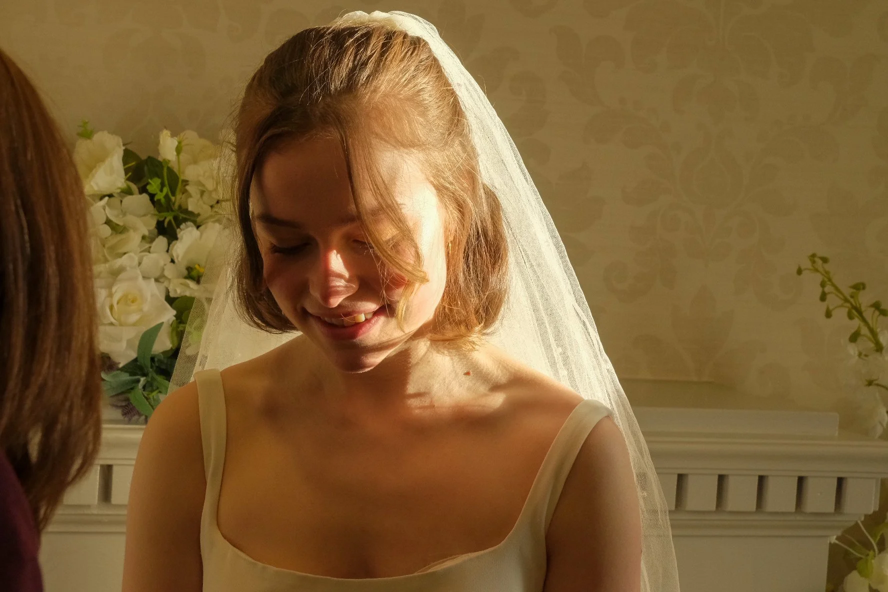 A smiling bride with a veil on her head, dressed in a cream-colored wedding gown, standing in front of a floral arrangement and ornate wallpaper.