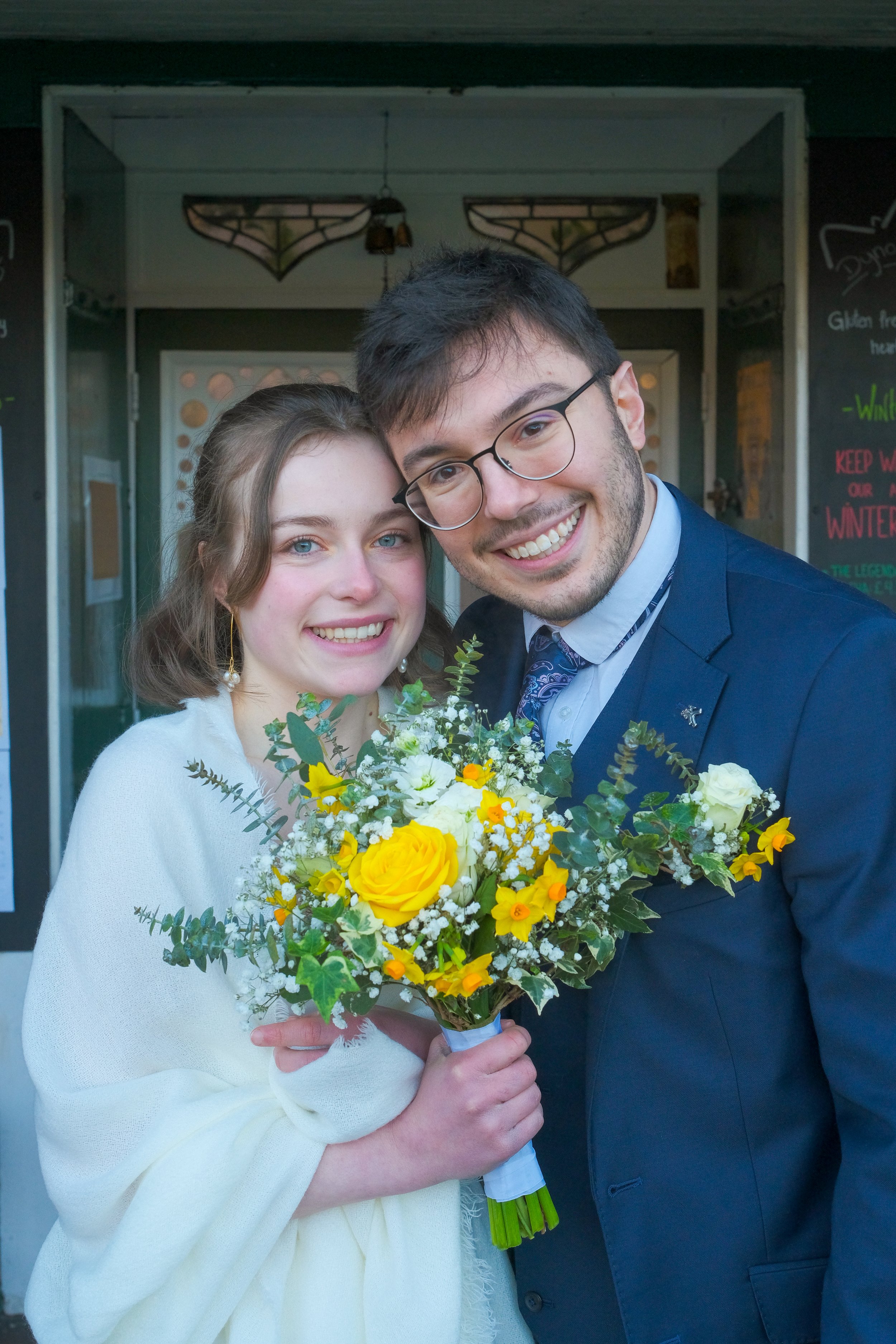 A happy couple smiling, with the woman holding a yellow and white floral bouquet, standing outside in front of a building with decorative lighting and a chalkboard sign.