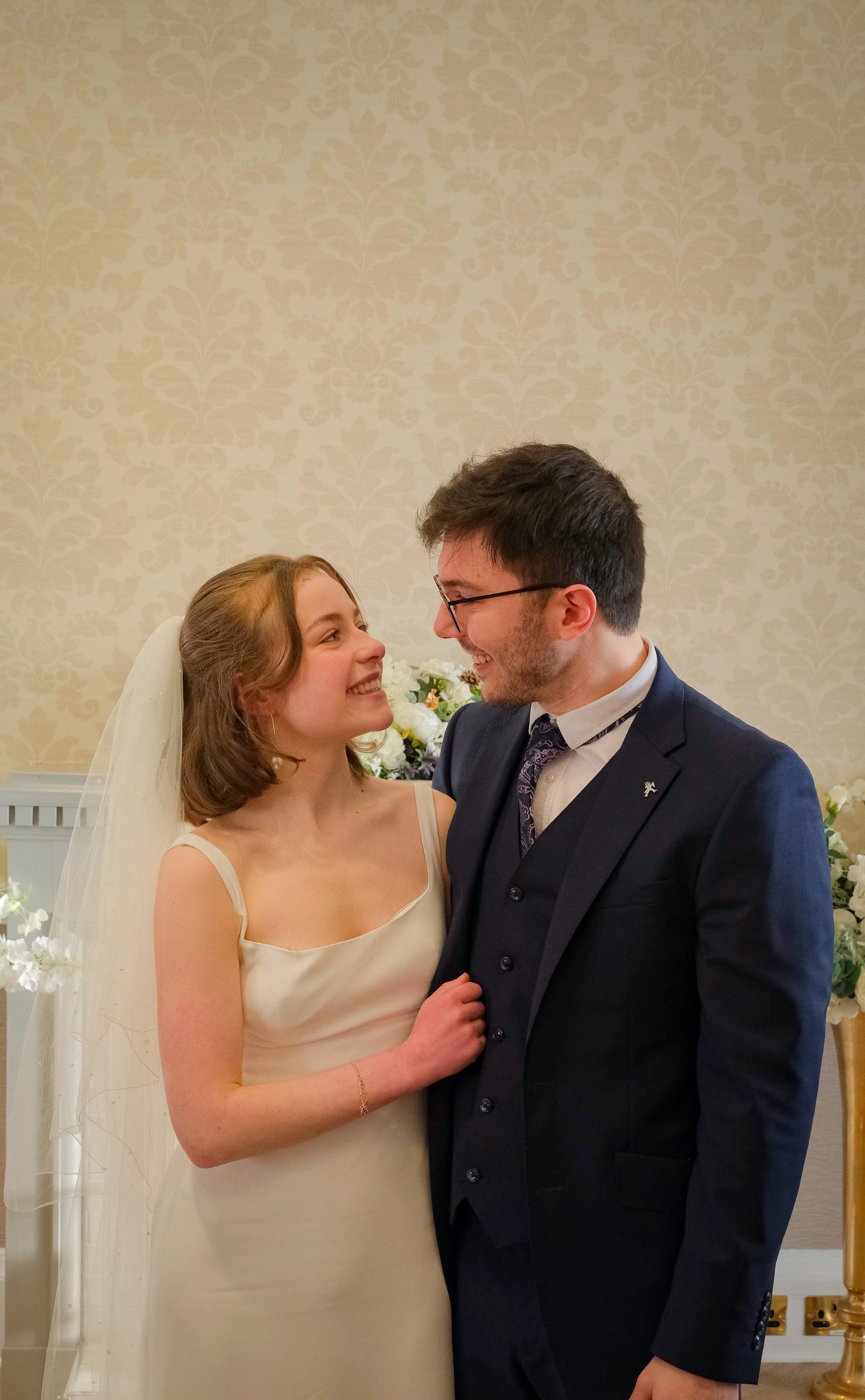 A bride and groom smiling and looking at each other indoors with floral decorations in the background.