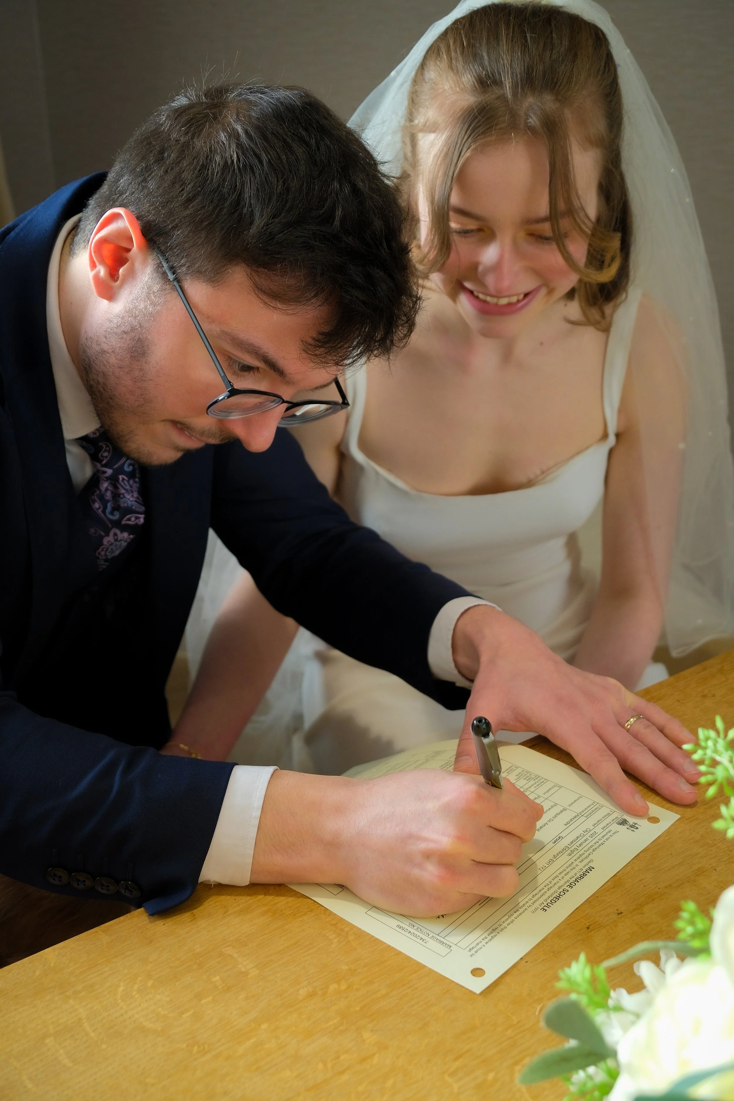 A young couple signing a marriage license at a wedding ceremony, with the bride smiling and leaning over the table.