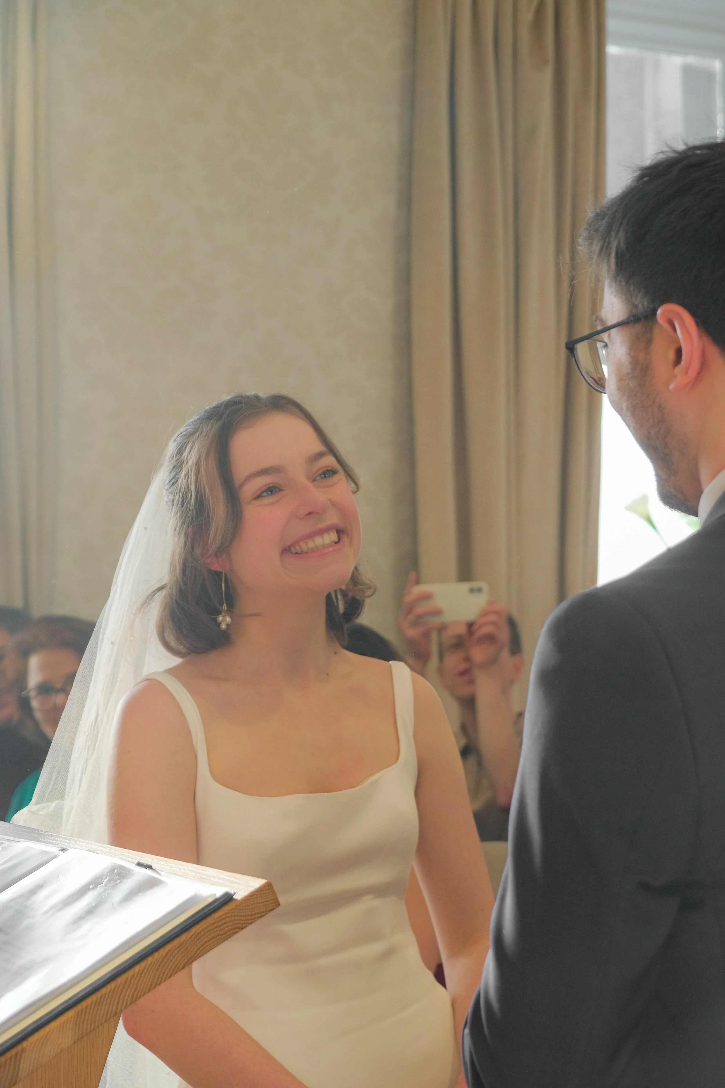 A smiling bride with shoulder-length brown hair, wearing a white sleeveless wedding dress and veil, looking at her groom during their wedding ceremony.