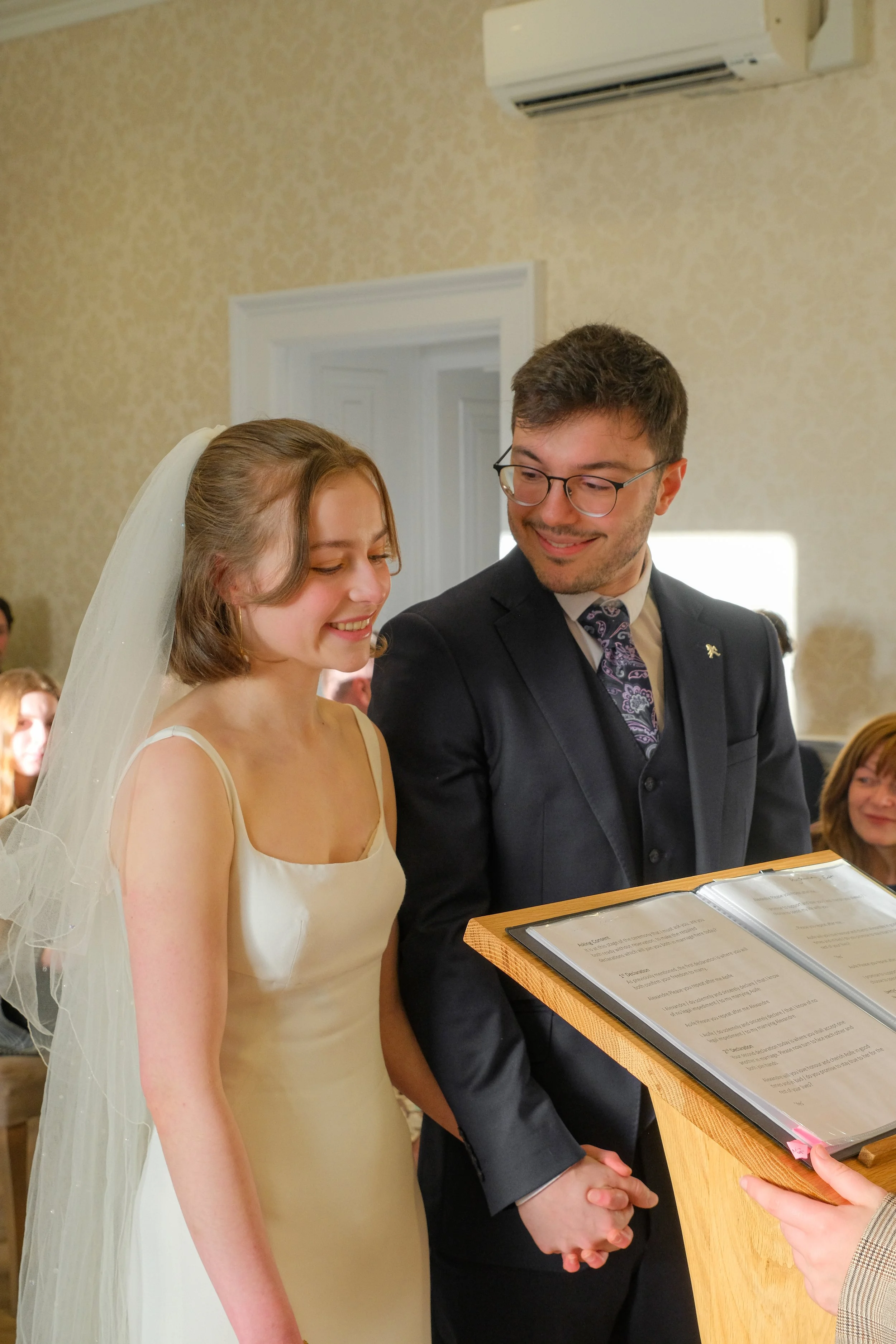 A bride and groom holding hands during their wedding ceremony, smiling at each other, with guests seated behind them.
