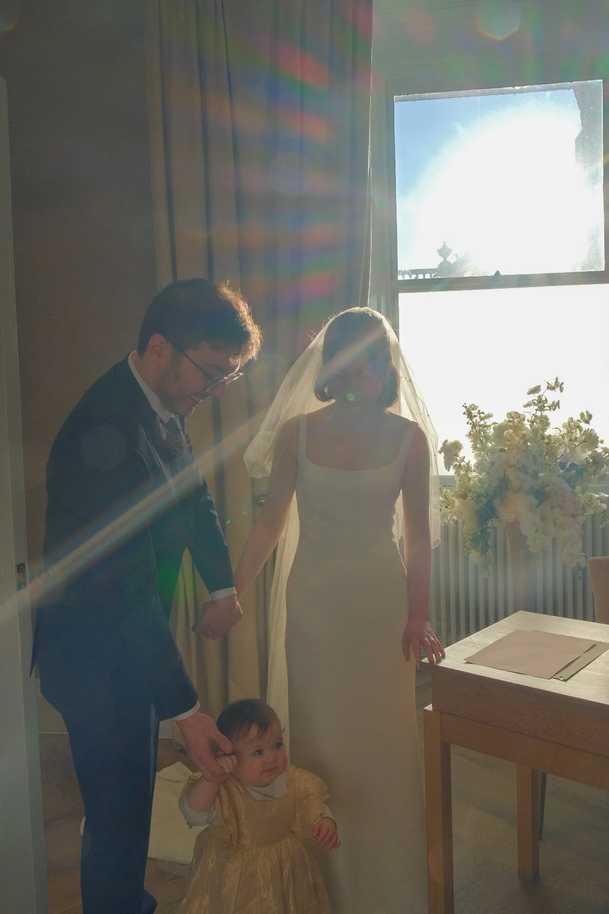 A bride and groom holding hands with a young child, possibly a flower girl, during a wedding ceremony inside a room with sunlight streaming through a window and flower arrangements on the side.