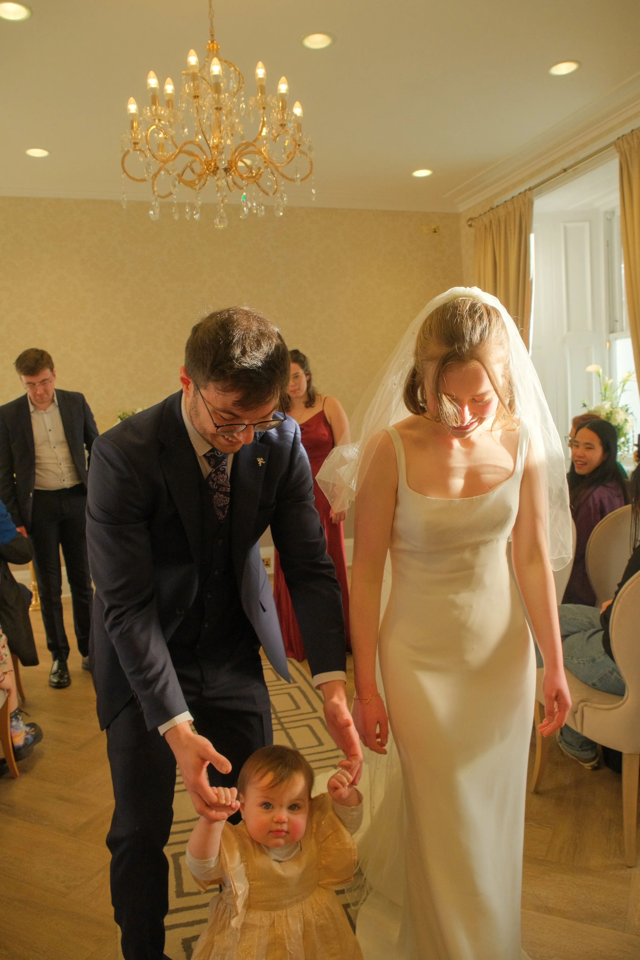 A bride and groom holding a child's hands during a wedding celebration in a decorated room with a chandelier and guests seated around.