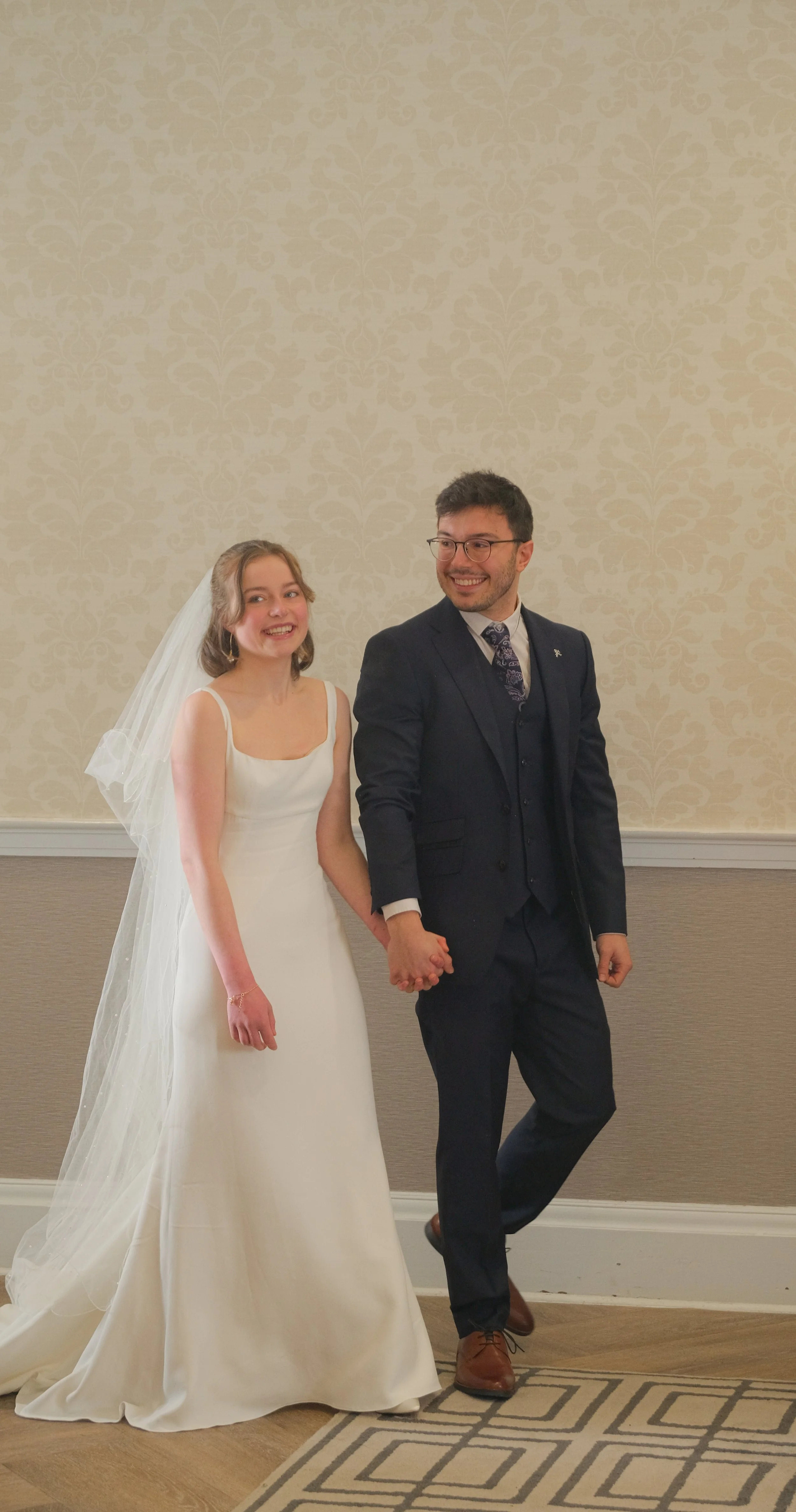 A bride and groom holding hands inside a wedding venue, smiling and looking at each other.