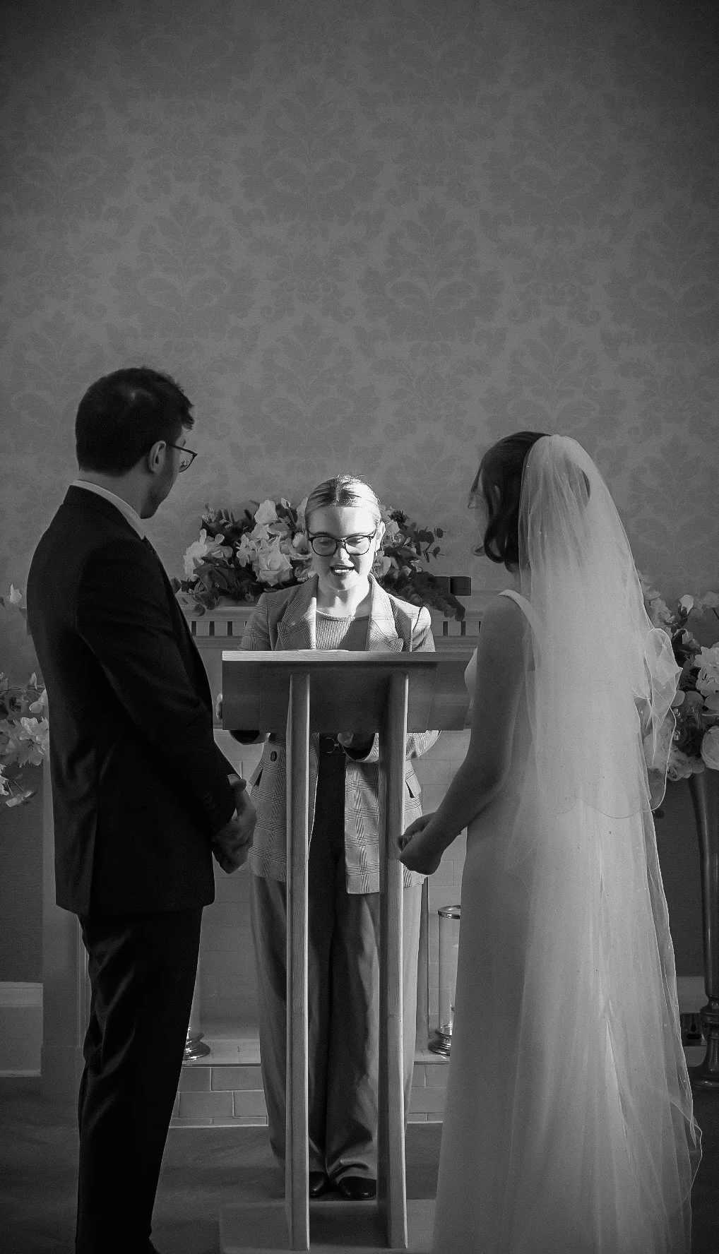A black and white photo of a couple getting married, standing before an officiant during a wedding ceremony. The groom is on the left wearing a suit, and the bride is on the right in a wedding dress and veil. The officiant is centered, smiling, holdi