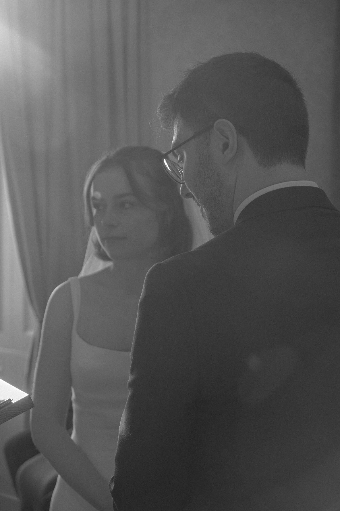 A black-and-white photo of a man in formal attire with glasses, and a woman in a wedding dress with a veil, indoors, standing near a window with curtains.