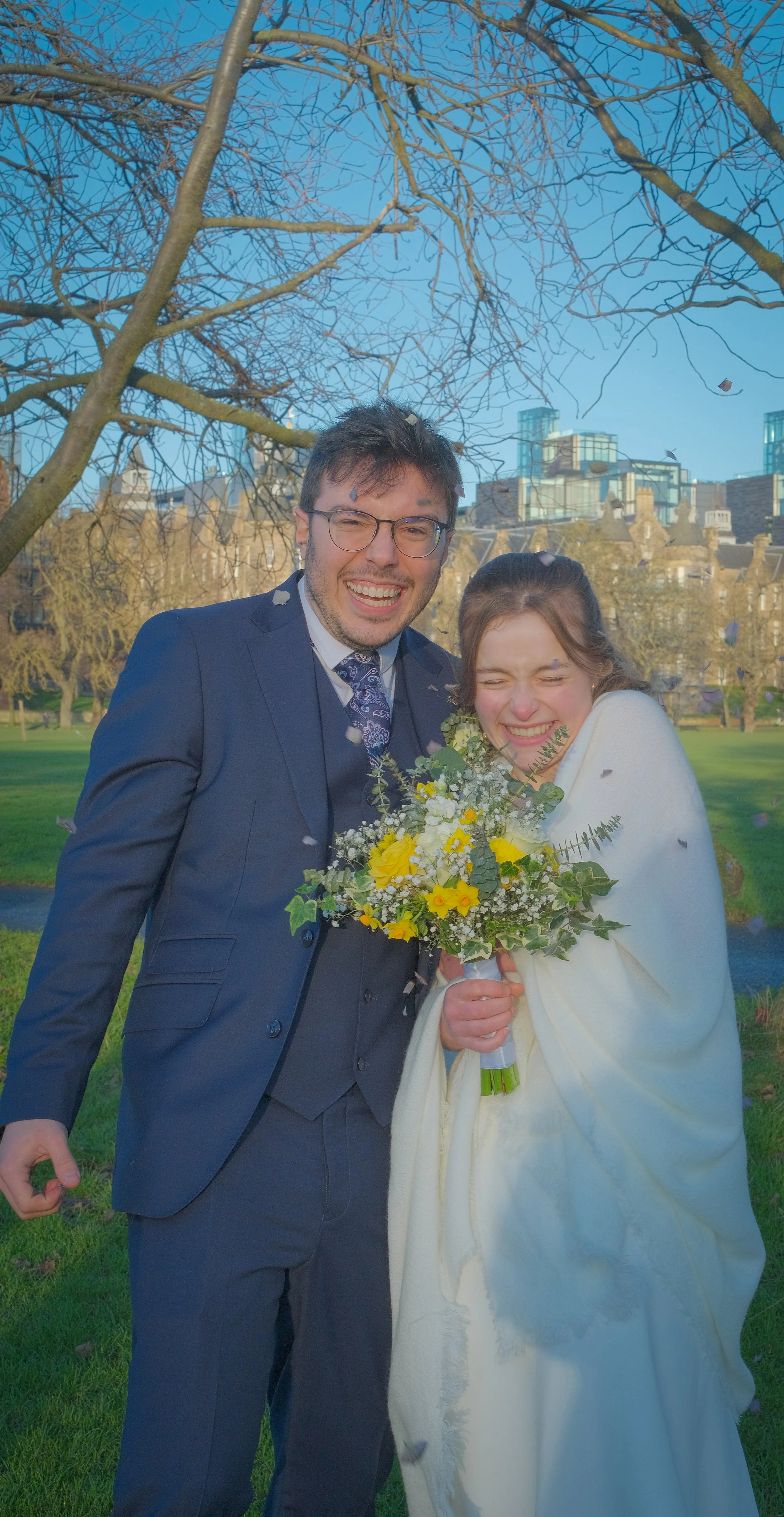 A happily married couple in wedding attire, smiling and celebrating outdoors with falling confetti, greenery, and city buildings in the background.