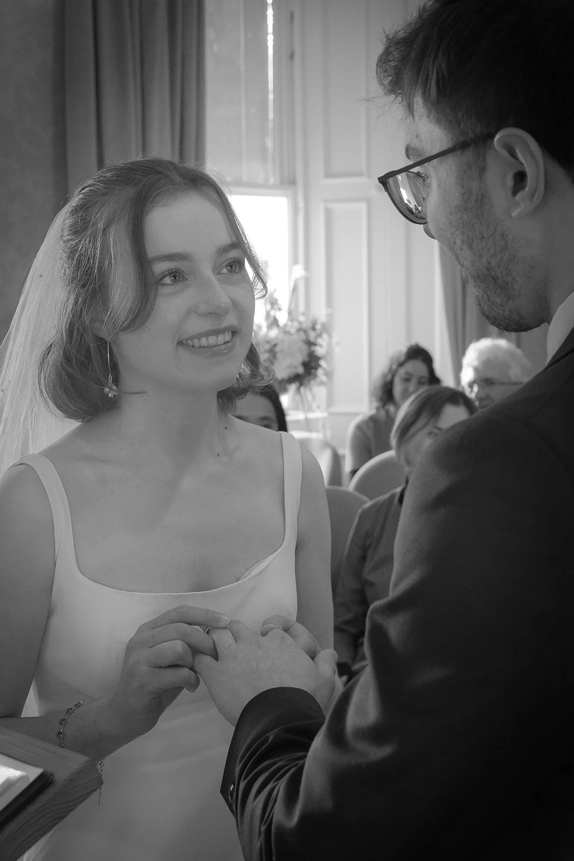 A bride and groom holding hands during their wedding ceremony, smiling and gazing at each other, in a room with guests and flowers in the background.