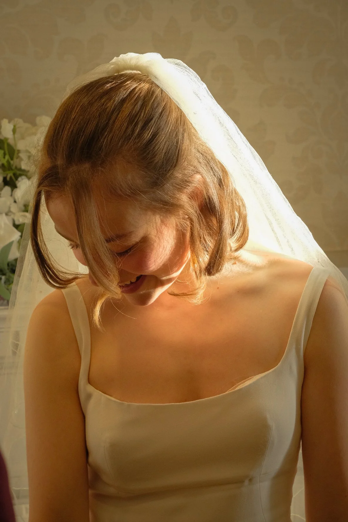 A bride looking downward, smiling, in a wedding dress with a veil, standing in front of a floral arrangement.