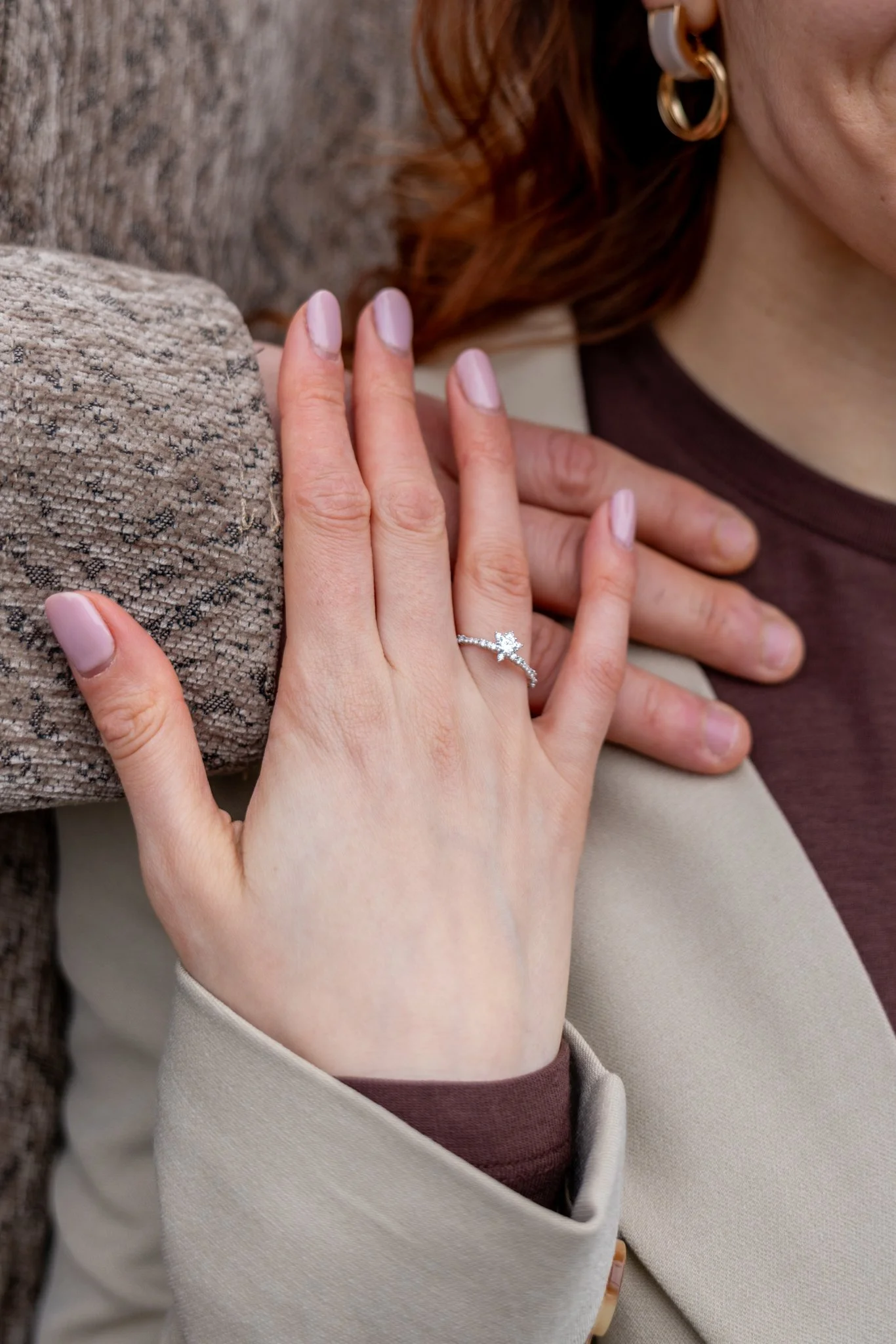 Close-up of a woman with red hair wearing gold hoop earrings and a beige blazer, resting her hand with a diamond ring on her finger on her chest, with a textured fabric surface in the background.