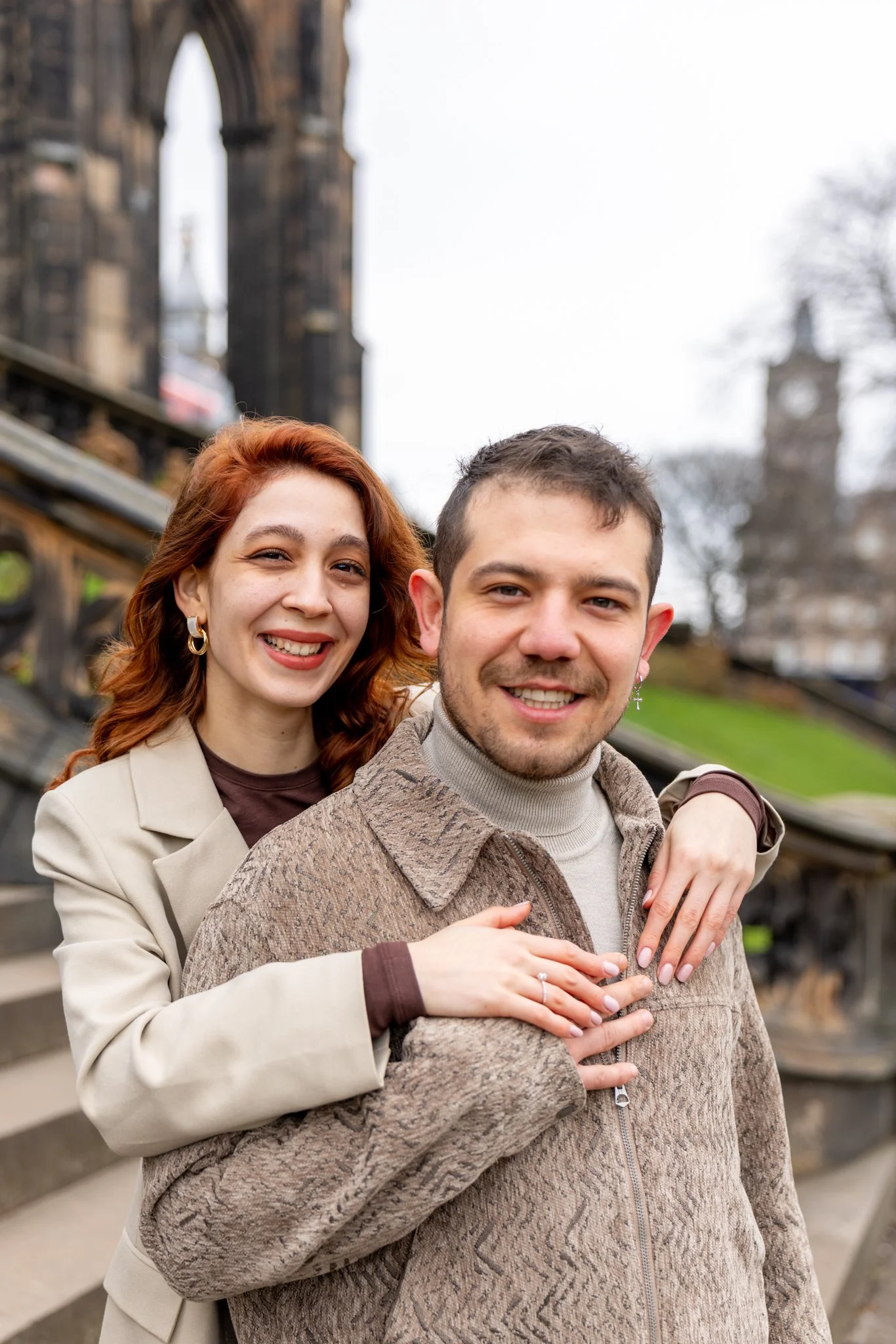 A smiling woman with red hair and a man with dark hair standing close together outdoors, with a historic stone building and clock tower in the background.
