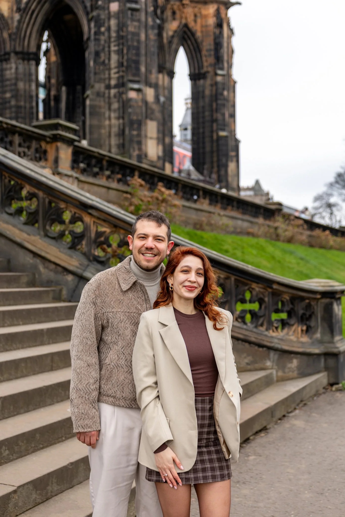 Two people smiling standing on stairs outdoors with historic Gothic-style building in the background.