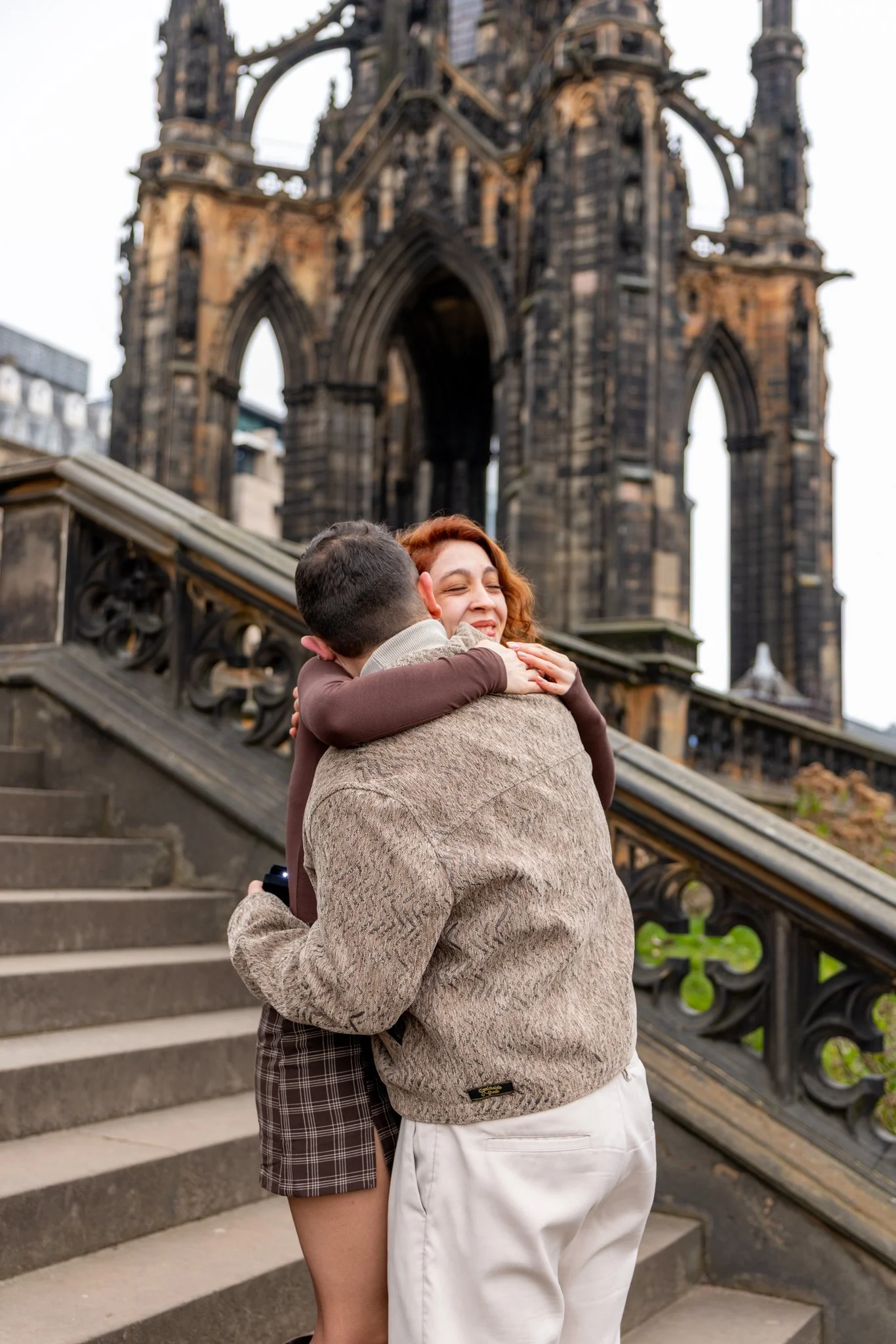 A man and a woman hugging on steps outside near a historical building with gothic architecture.
