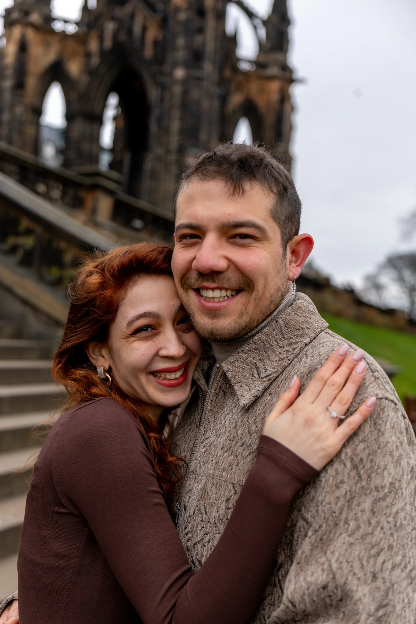 A happy couple hugging each other outdoors with a historic, gothic-style building in ruins in the background.