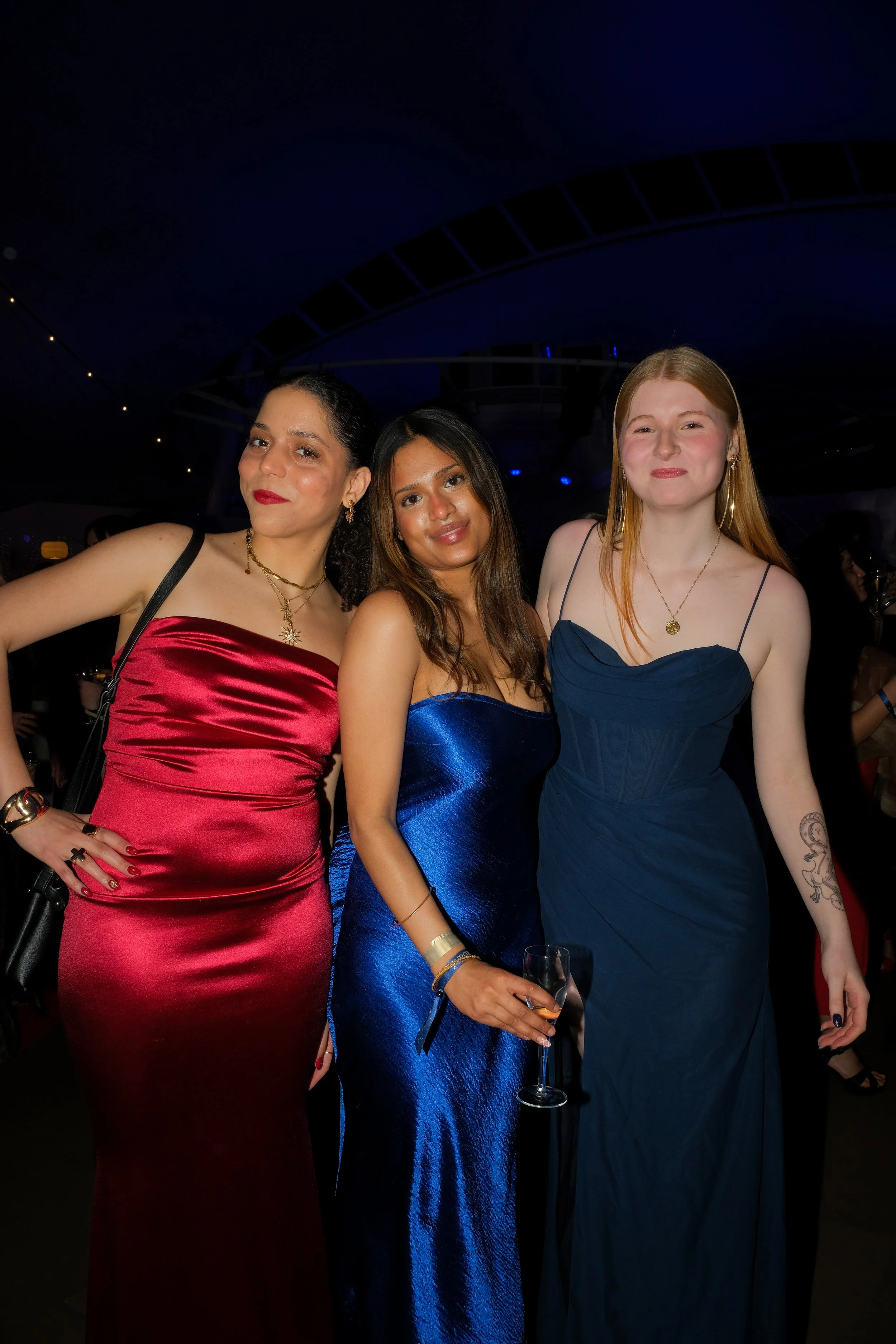 Three women at a party wearing elegant dresses, with one holding a glass of champagne, standing close together and smiling.