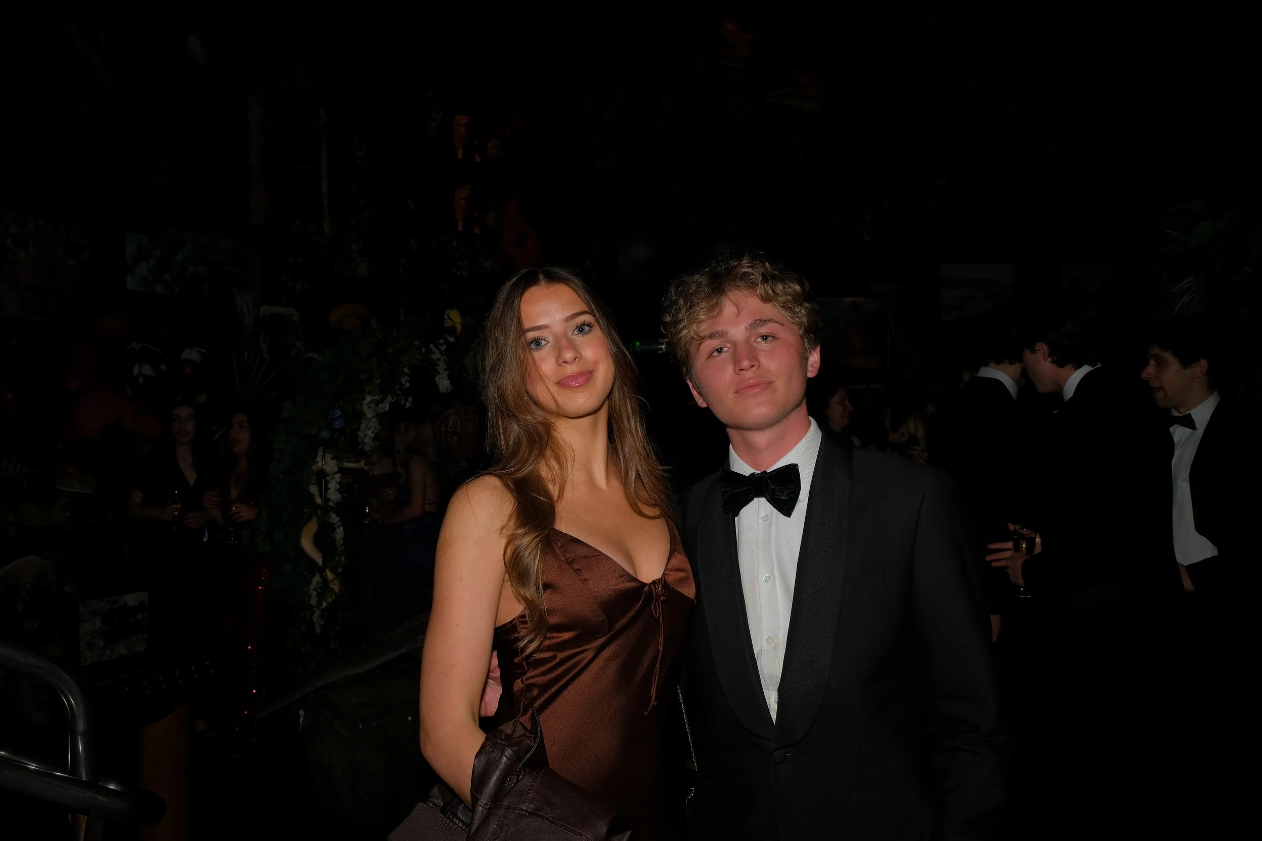 A young woman in a brown satin dress and a young man in a black tuxedo with a bow tie posing together at a formal event.