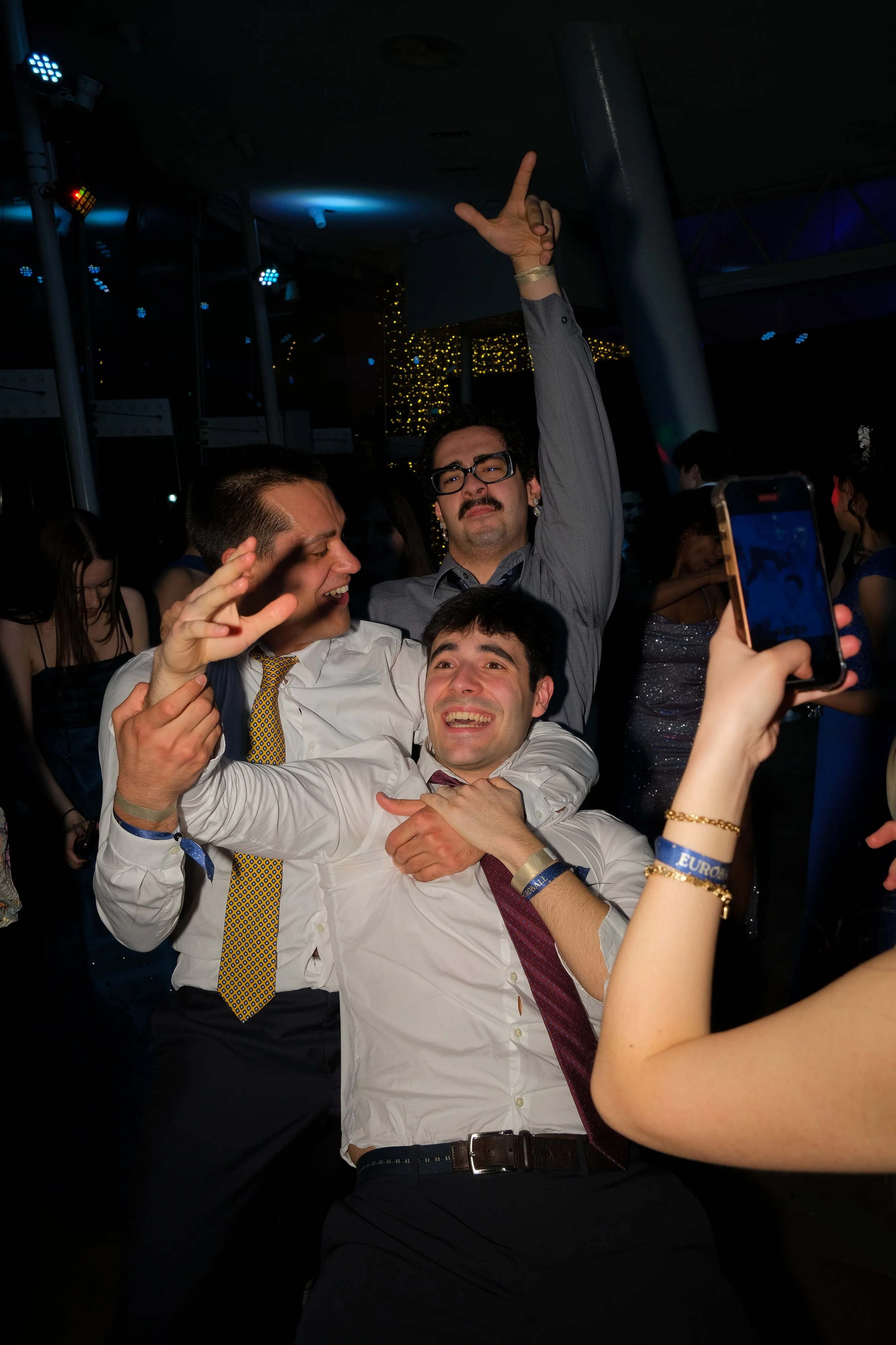 Group of men celebrating and taking photos at a lively party or wedding reception with dark lighting and festive decorations.