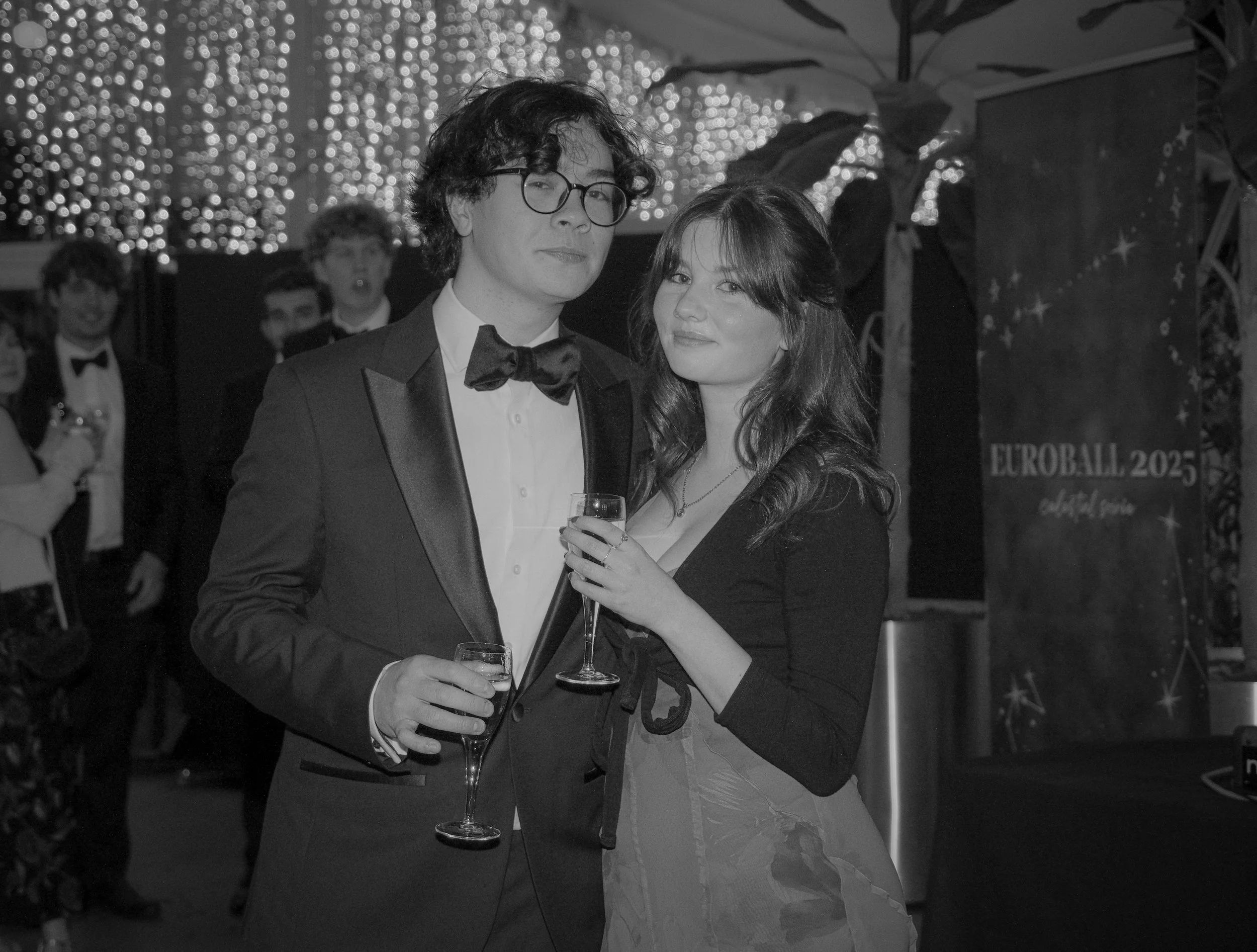 A black and white photo of a man in a tuxedo with a bow tie and glasses, and a woman in a black top with a necklace and a skirt, both holding champagne glasses, at a formal event with a sign in the background reading "EUROBALL 2025" and other guests 
