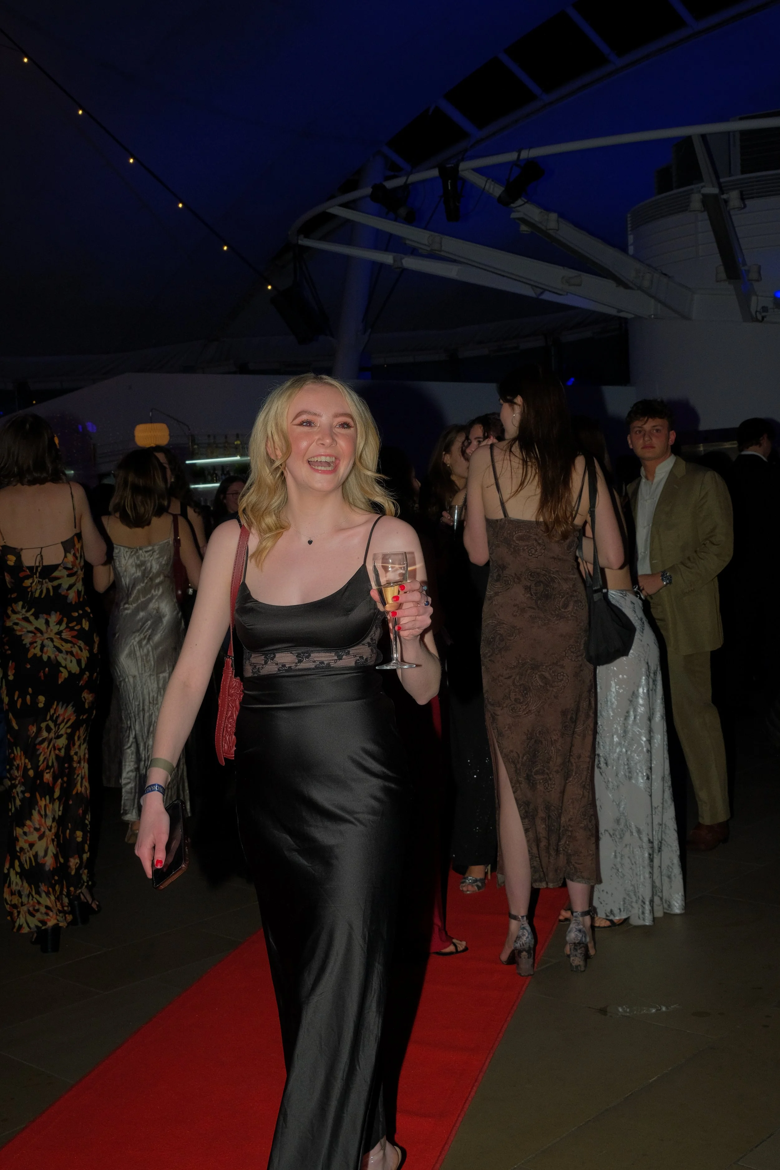 A young woman in a black satin gown holding a glass of sparkling wine at a party on a red carpet, smiling at the camera, with other elegantly dressed guests in the background under a dark evening sky.