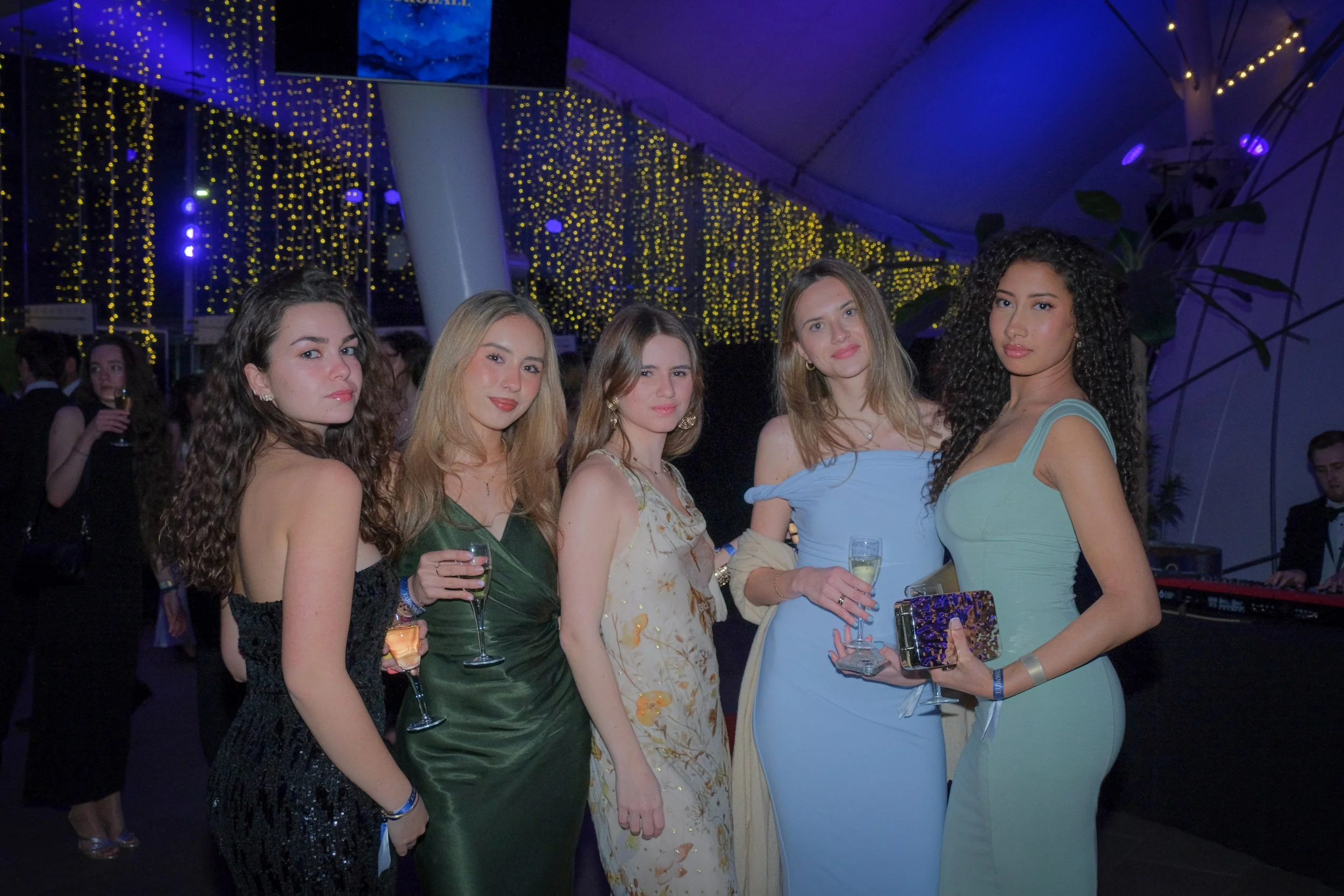Six women dressed in elegant gowns holding drinks at a party or event with decorative string lights in the background.