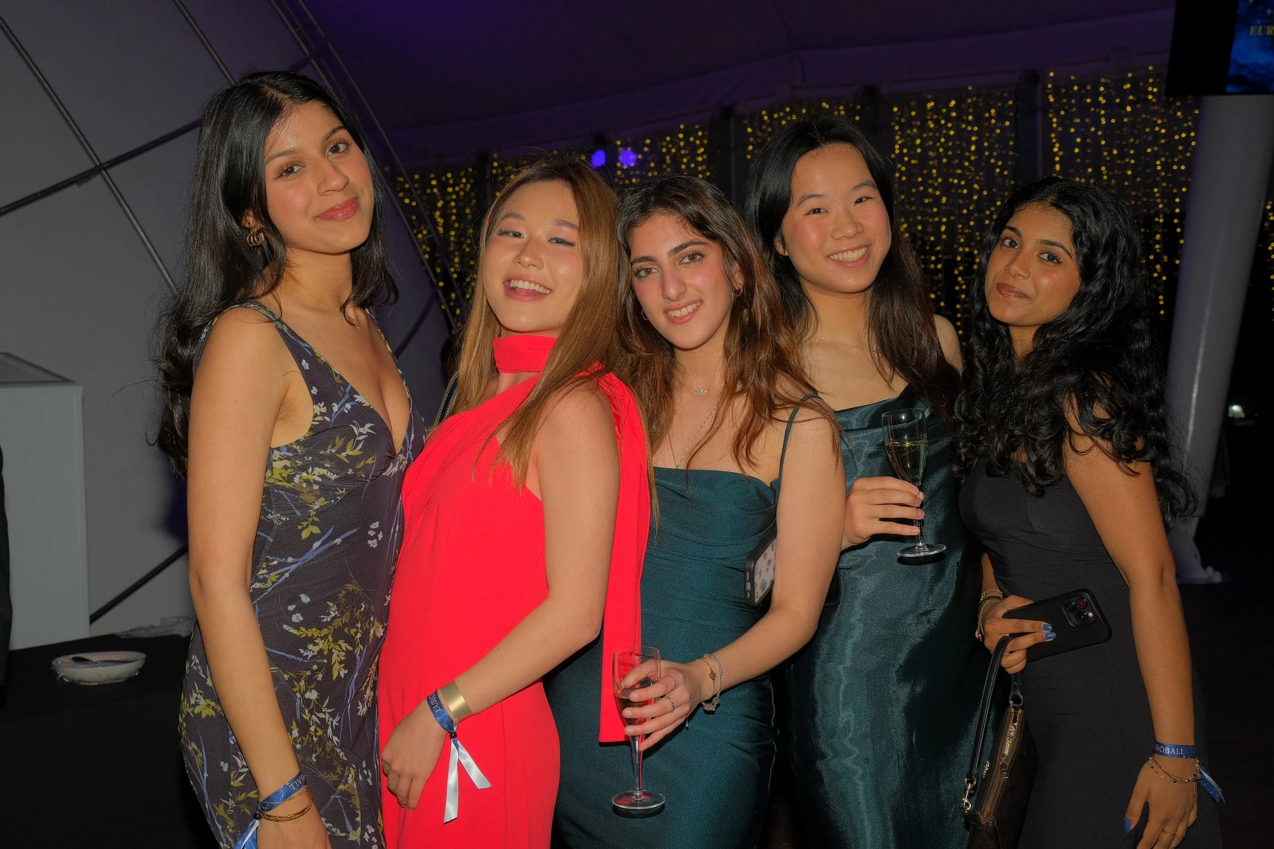 Group of five women dressed in elegant evening wear at a celebration or party, holding drinks, with festive lighting in the background.