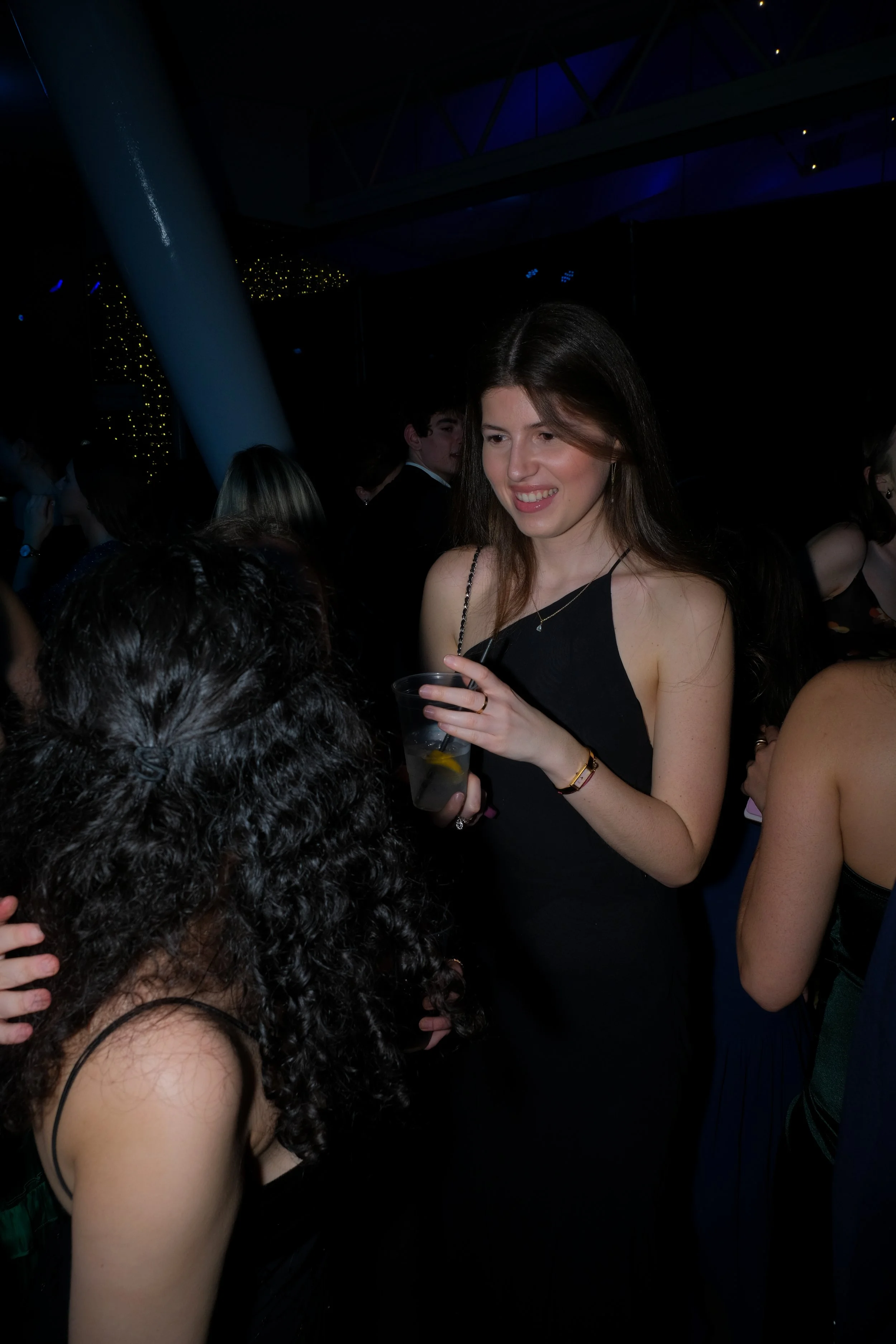 A young woman in a black dress holding a drink, smiling and talking to another person at a crowded party or event at night.