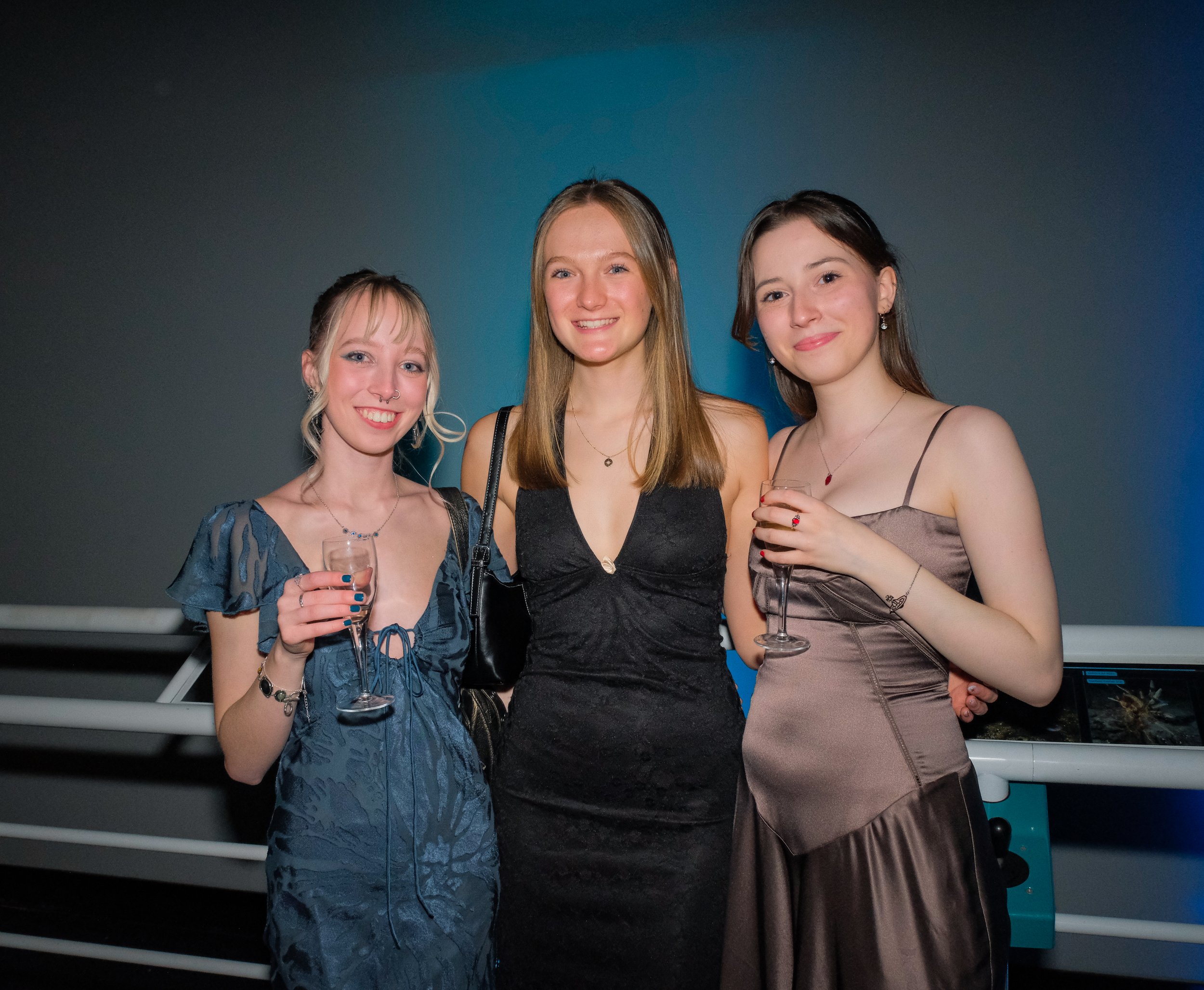 Three young women in formal dresses holding glasses of champagne at a social event, standing close together and smiling at the camera.