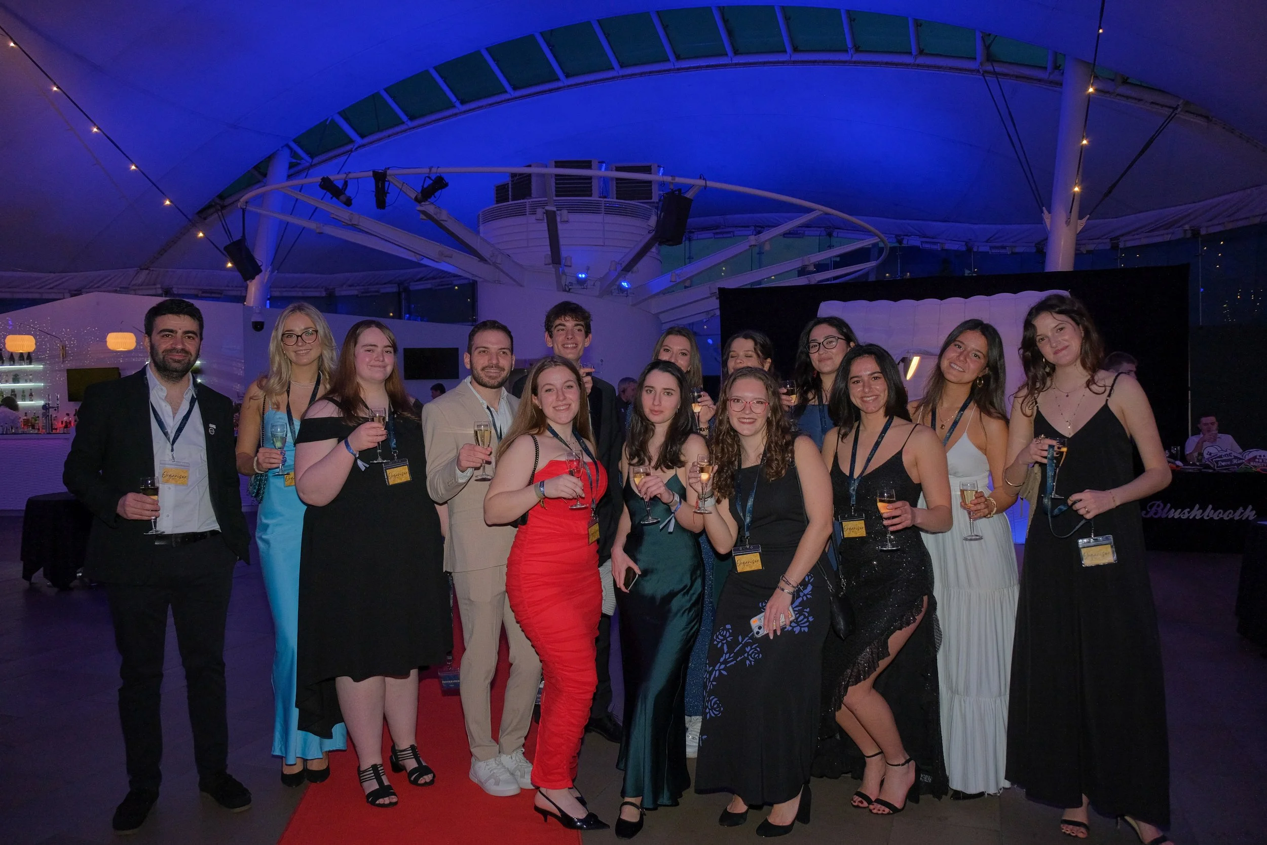 Group of young adults dressed in formal attire, celebrating with drinks at a social event or party in an indoor venue with blue lighting and modern decor.