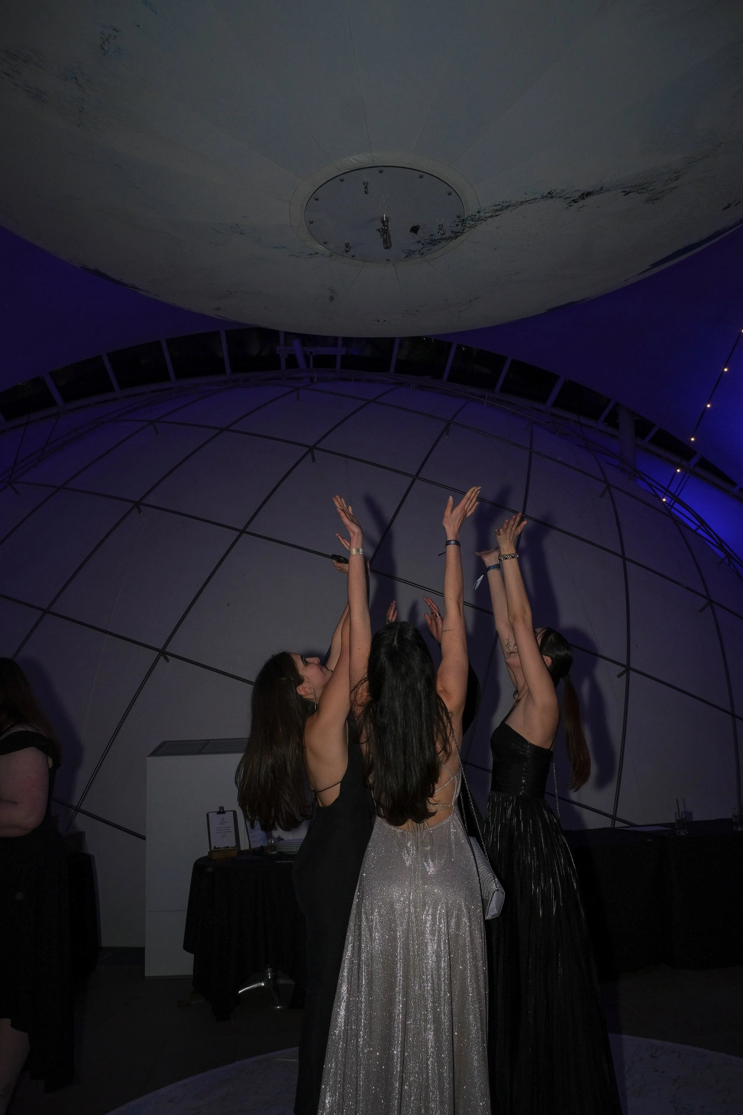 Three women in evening dresses reaching up towards the ceiling of a modern, dome-shaped building during an event.