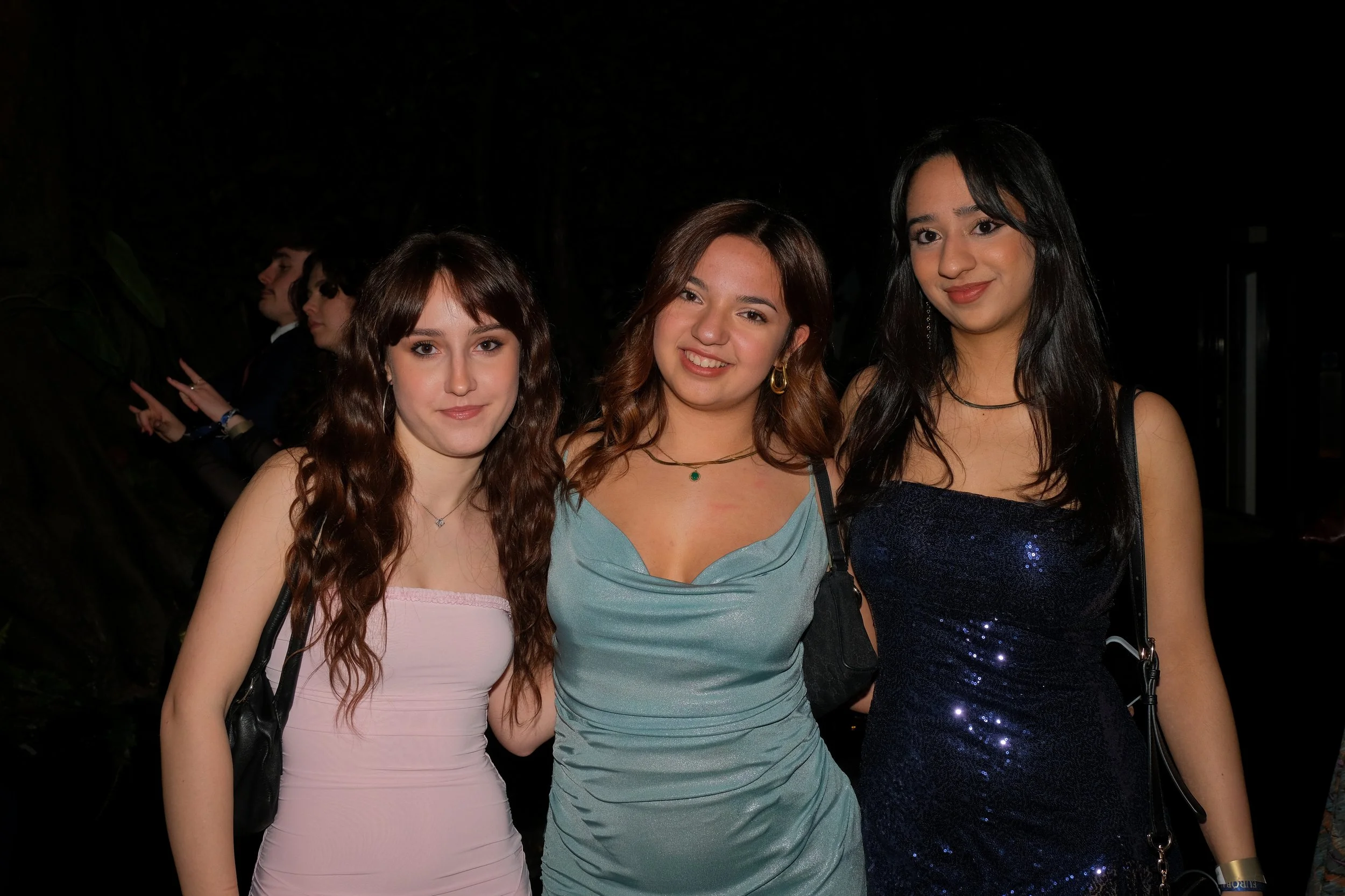 Three young women standing close together at a nighttime event, all smiling and posing for the camera, with two men in the background making peace signs.