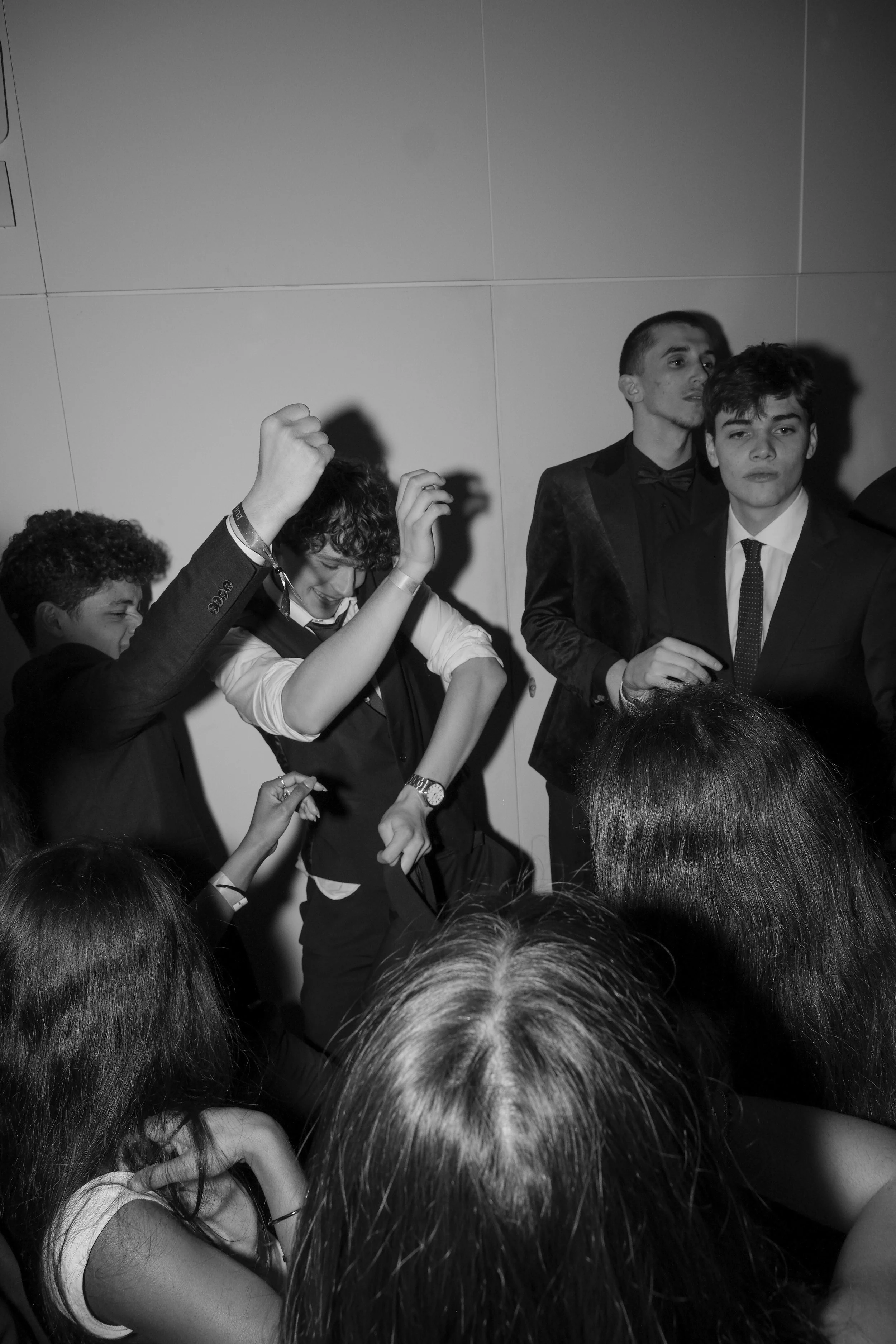 Black and white photo of young people at a party or event, some dancing and others watching, with a plain wall in the background.