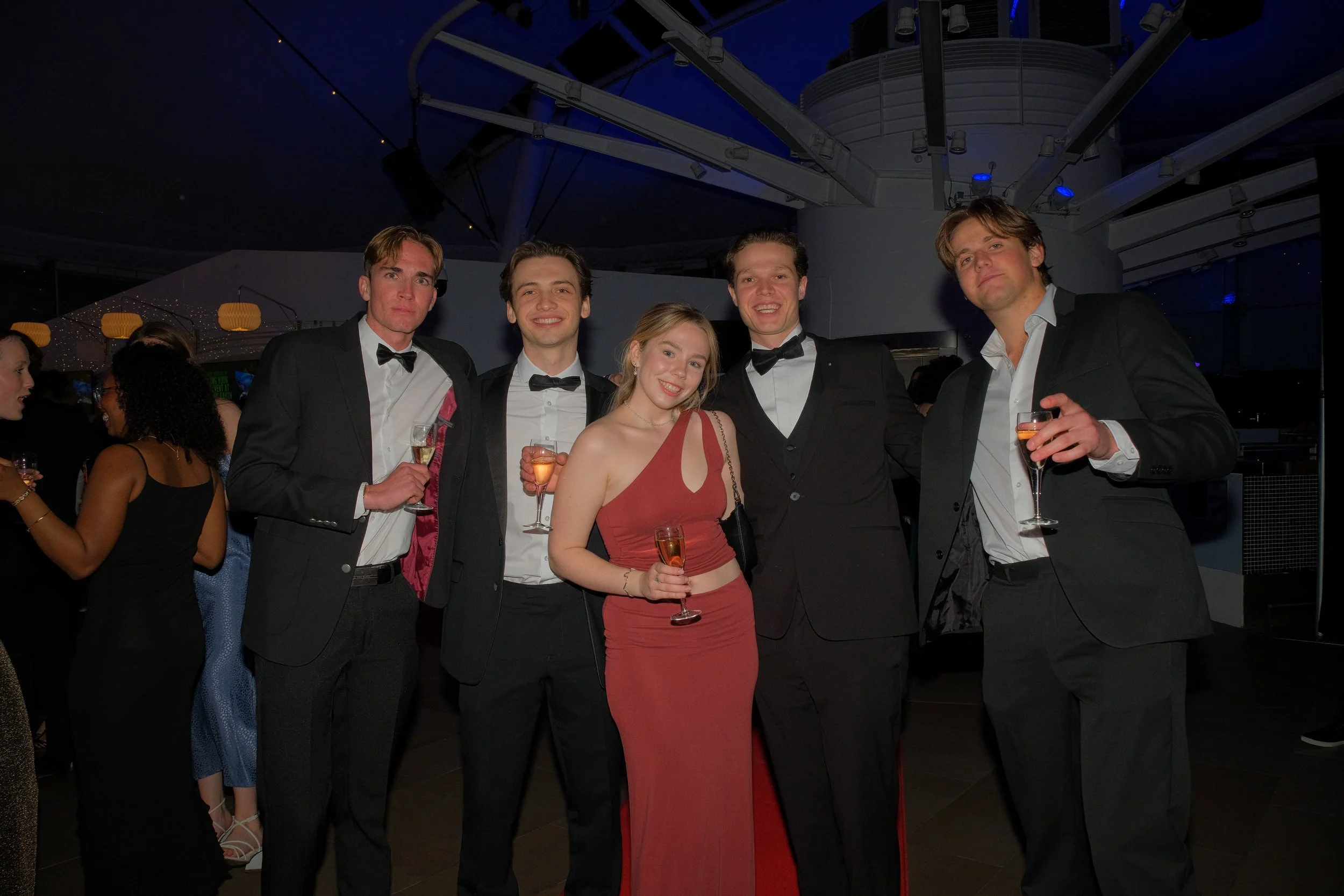 Group of five young adults dressed in formal attire, holding champagne glasses at a party or celebration.