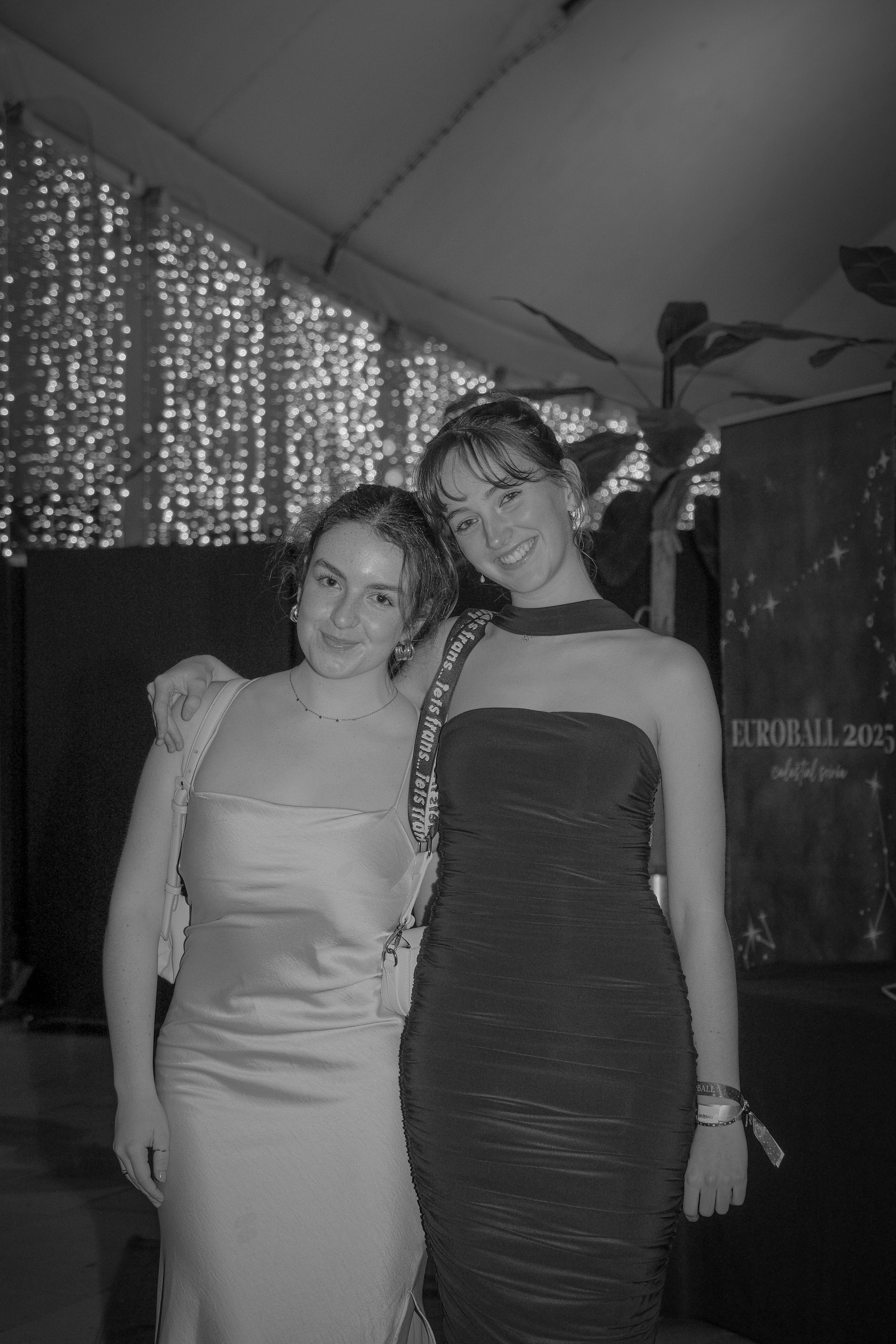 Two young women in elegant dresses smiling at a party with string lights in the background.