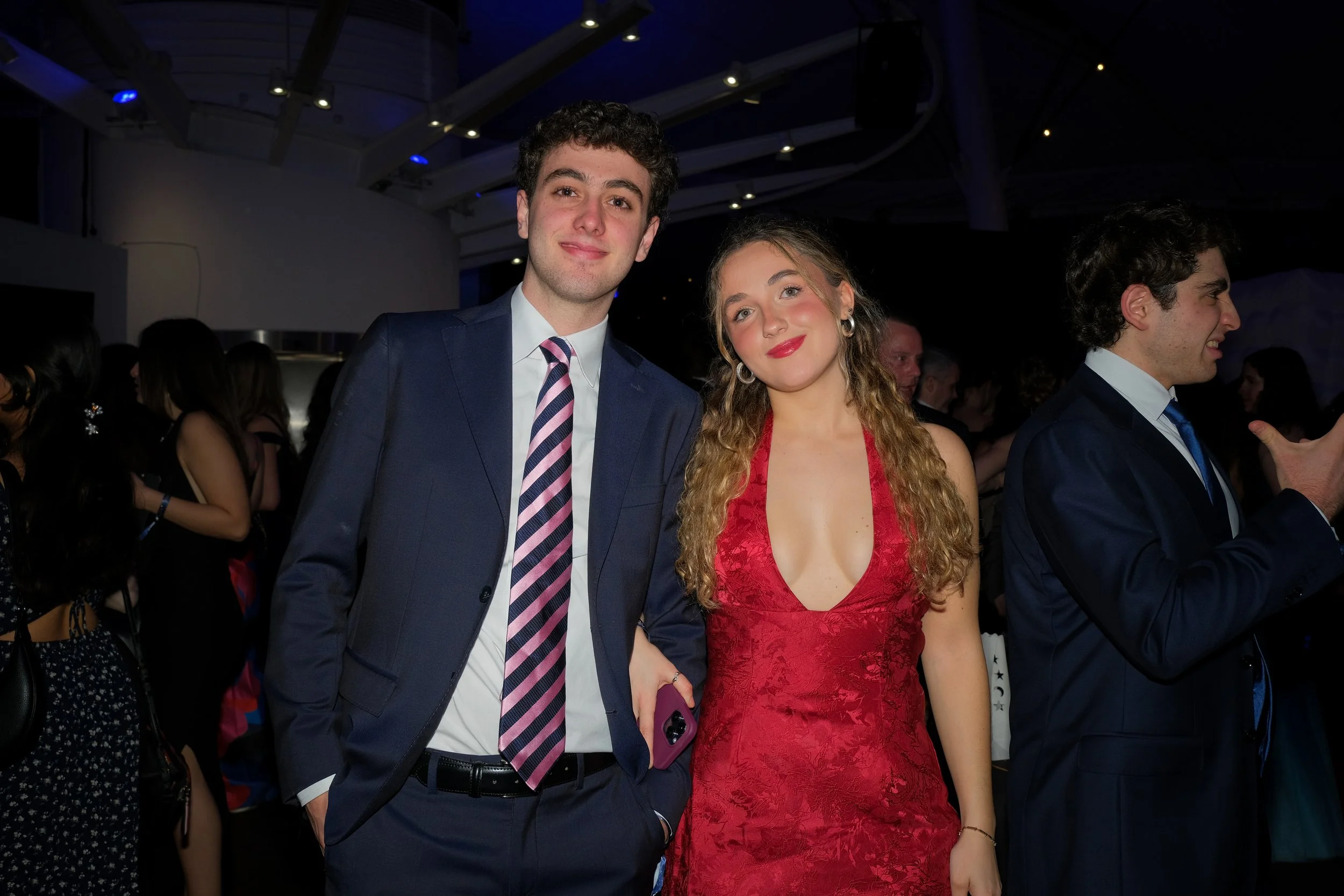A young man in a navy suit and striped tie standing next to a young woman in a red dress, both smiling at the camera at a formal event.
