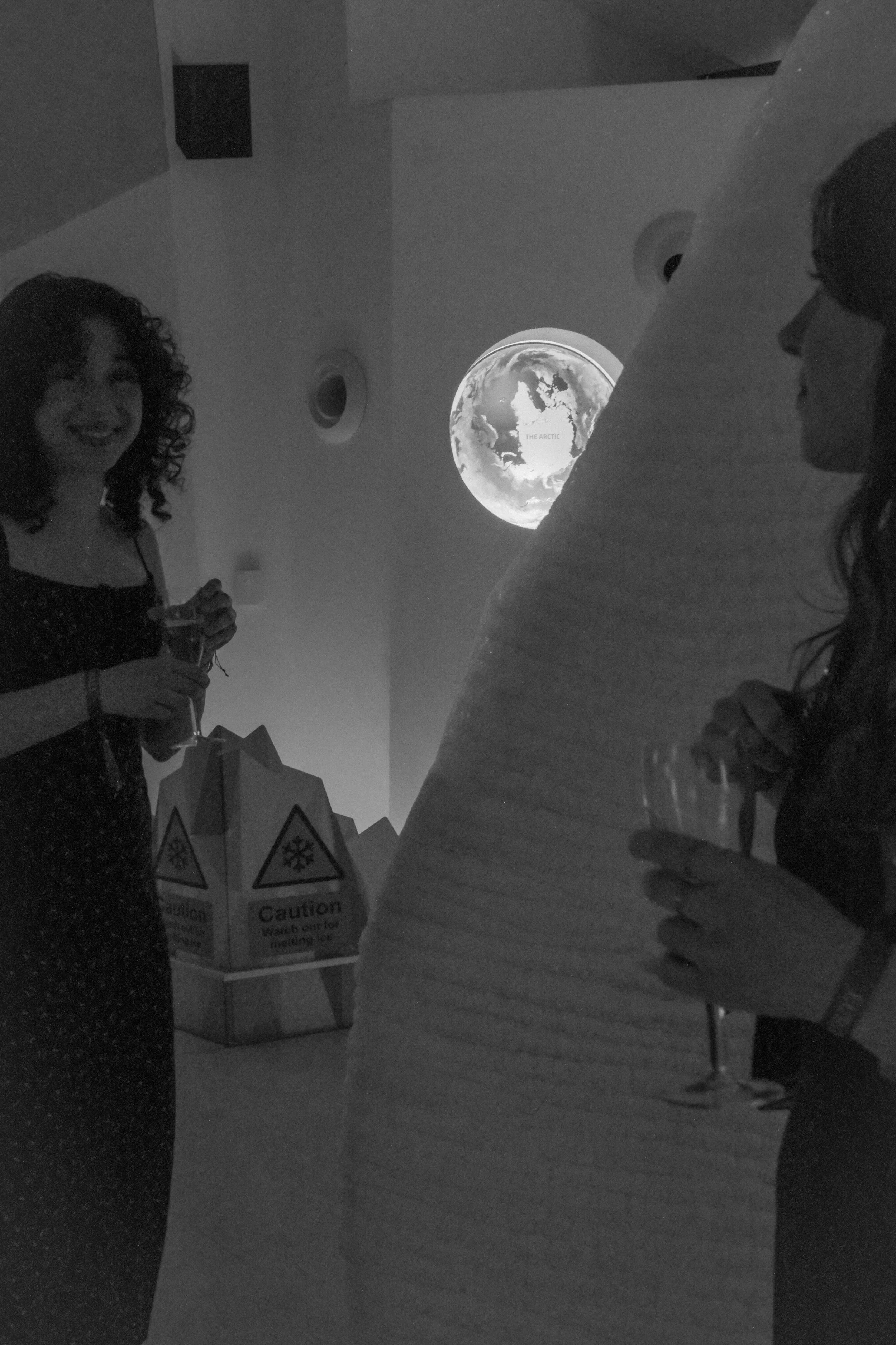 Two women talking at a party, one holding a drink, with a projected image of Earth in the background.