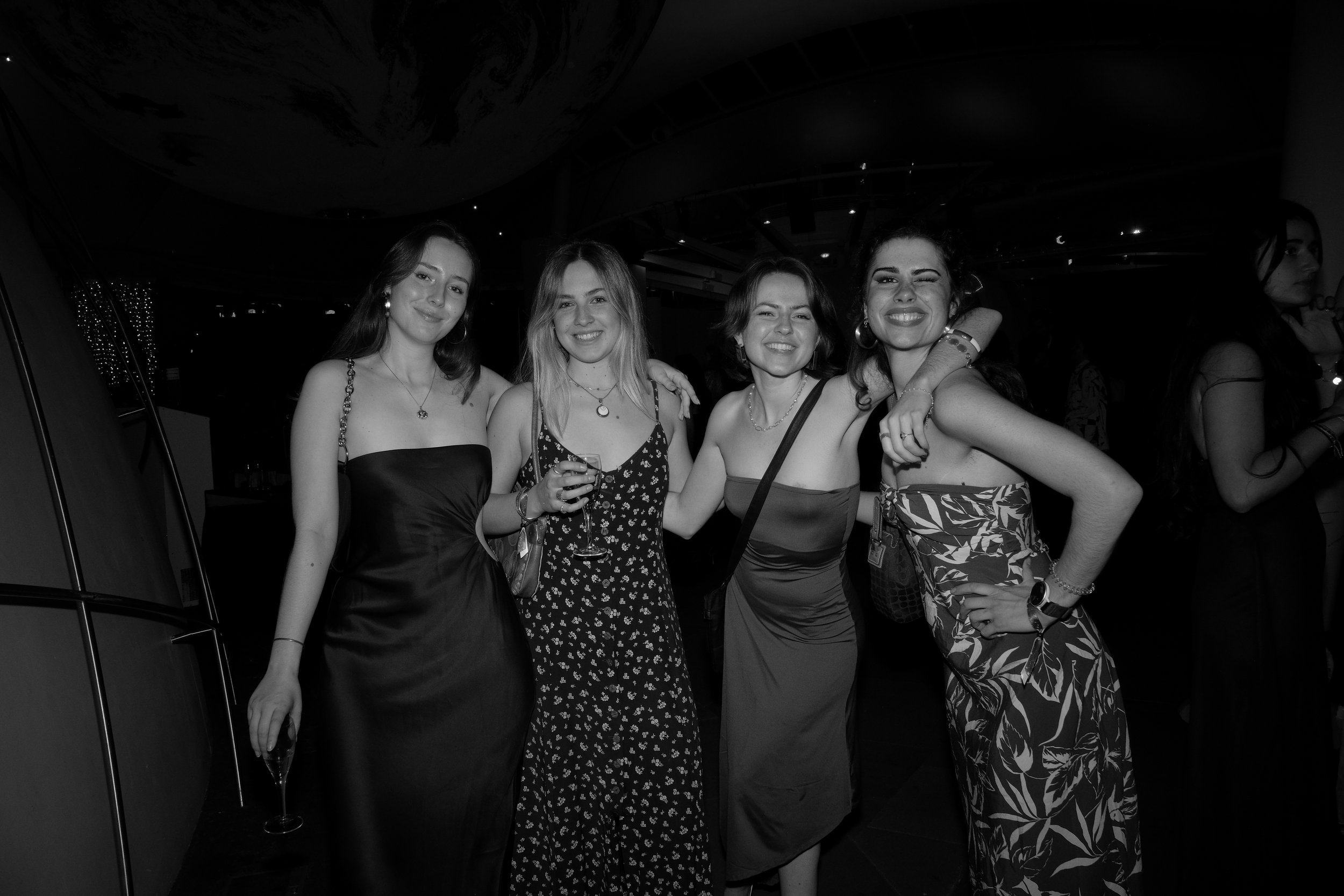 Four women in dresses smiling and posing together at a social event, with one holding a glass of wine.