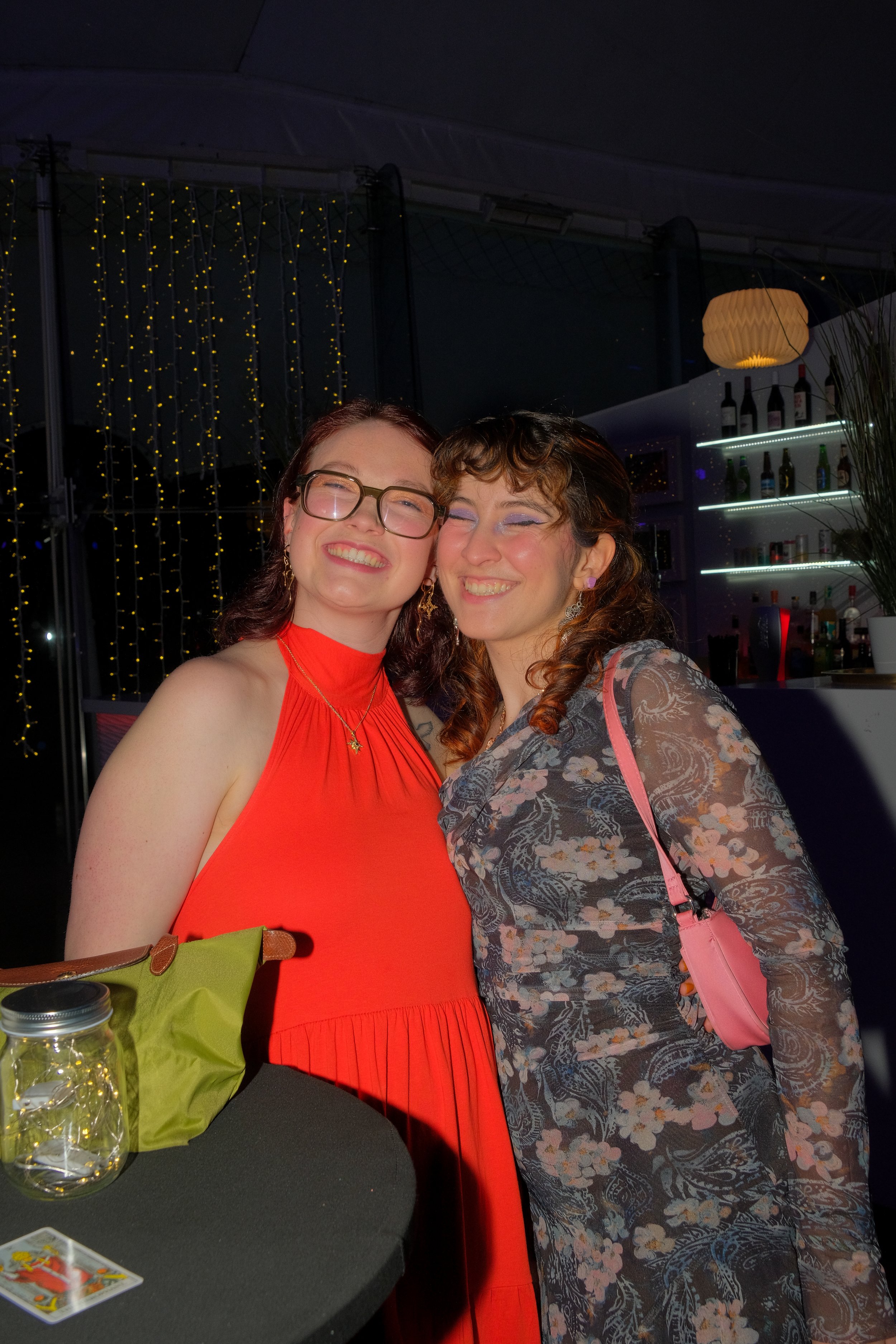 Two women smiling and posing closely together at a social event or party, with a bar and decorative string lights in the background.