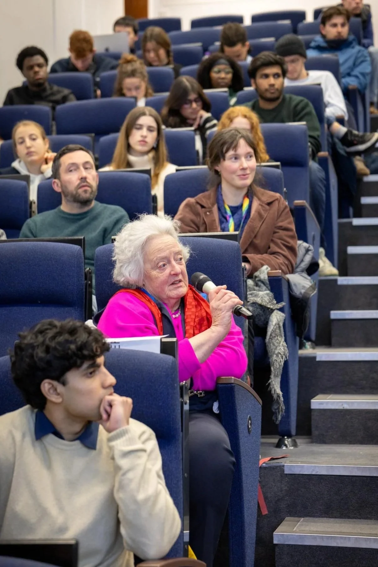 An older woman with white hair, wearing a pink jacket, sitting in an auditorium and speaking into a microphone, surrounded by an audience of diverse individuals.