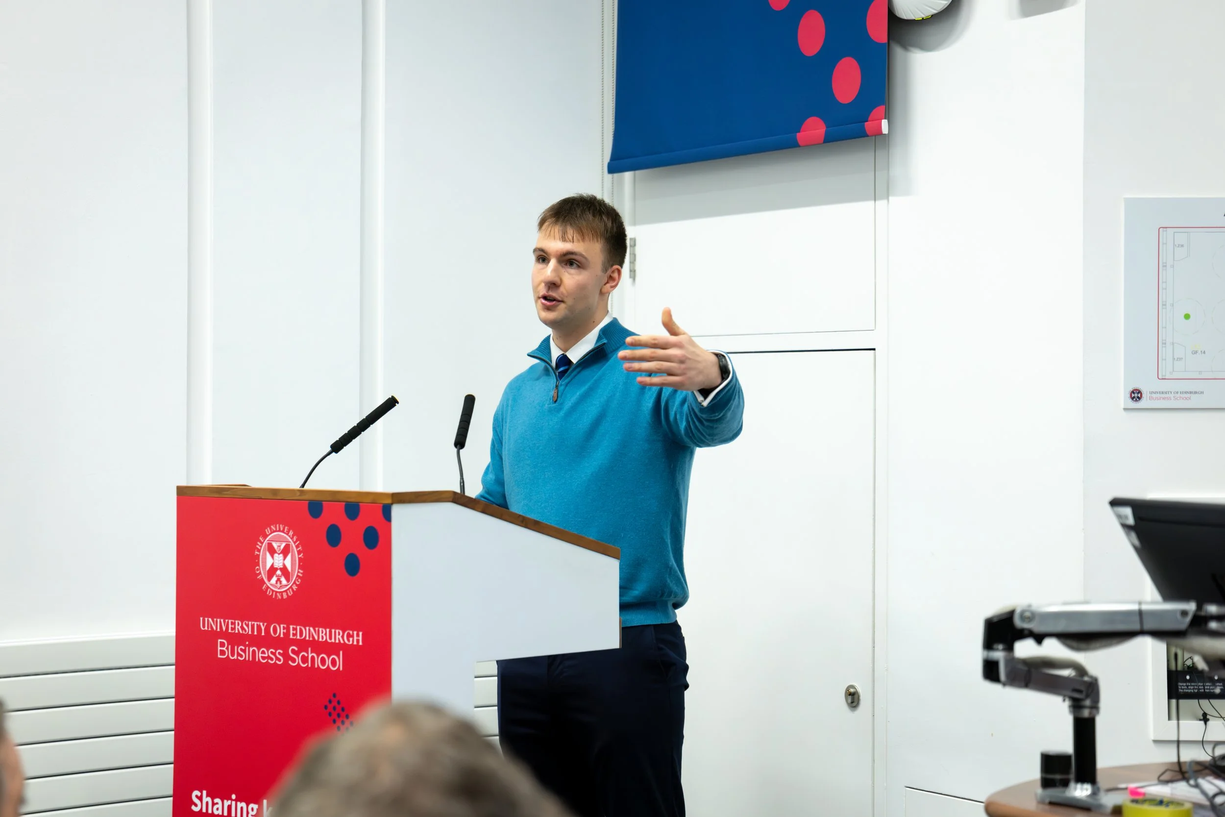A man in a blue sweater and dark pants giving a presentation at the University of Edinburgh Business School, standing behind a red podium with the university's logo, with a projector in the foreground and a white wall with a poster of a hockey rink i