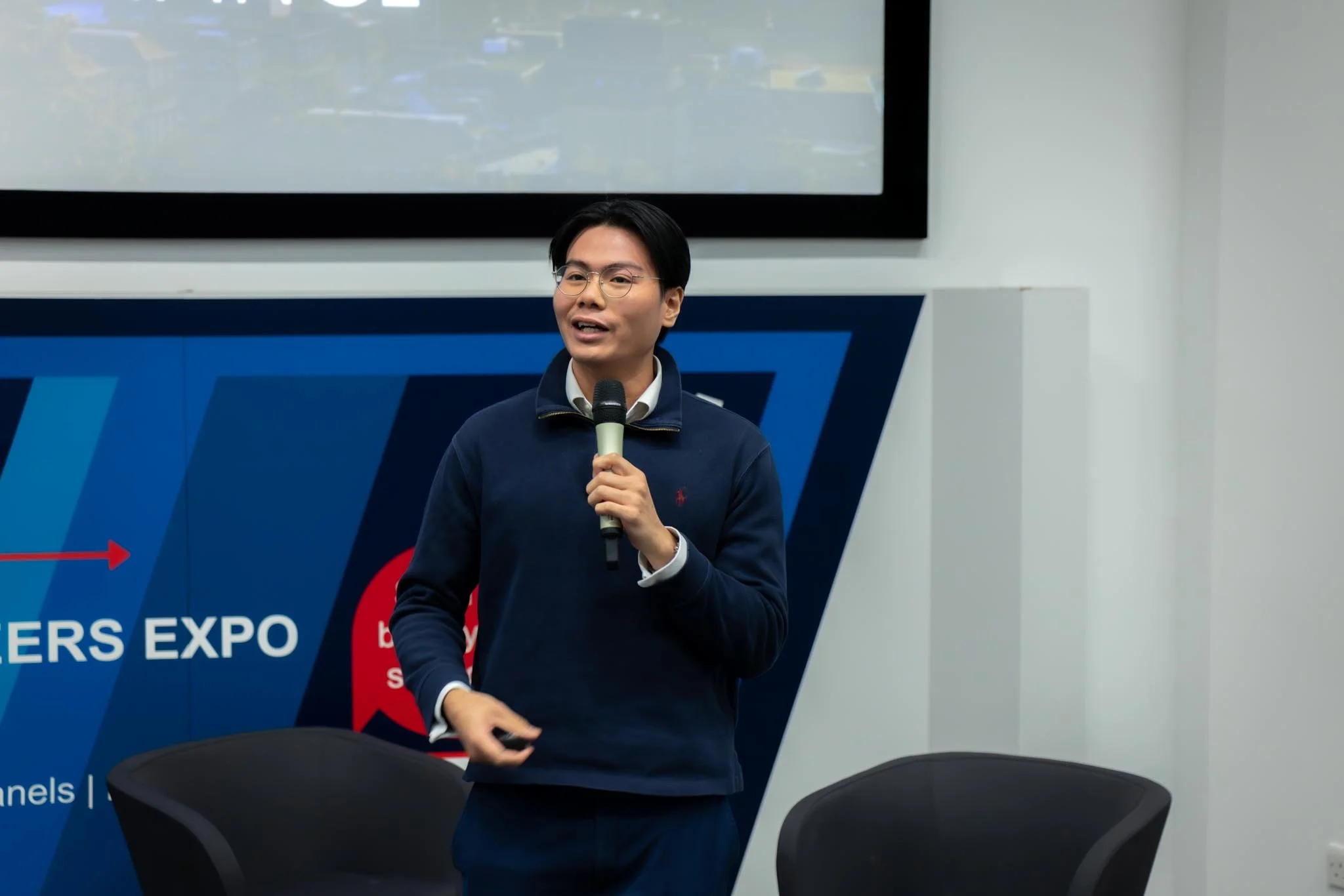 A young man in glasses and a navy blue sweater holding a microphone, speaking at an expo or conference in a classroom or conference room.