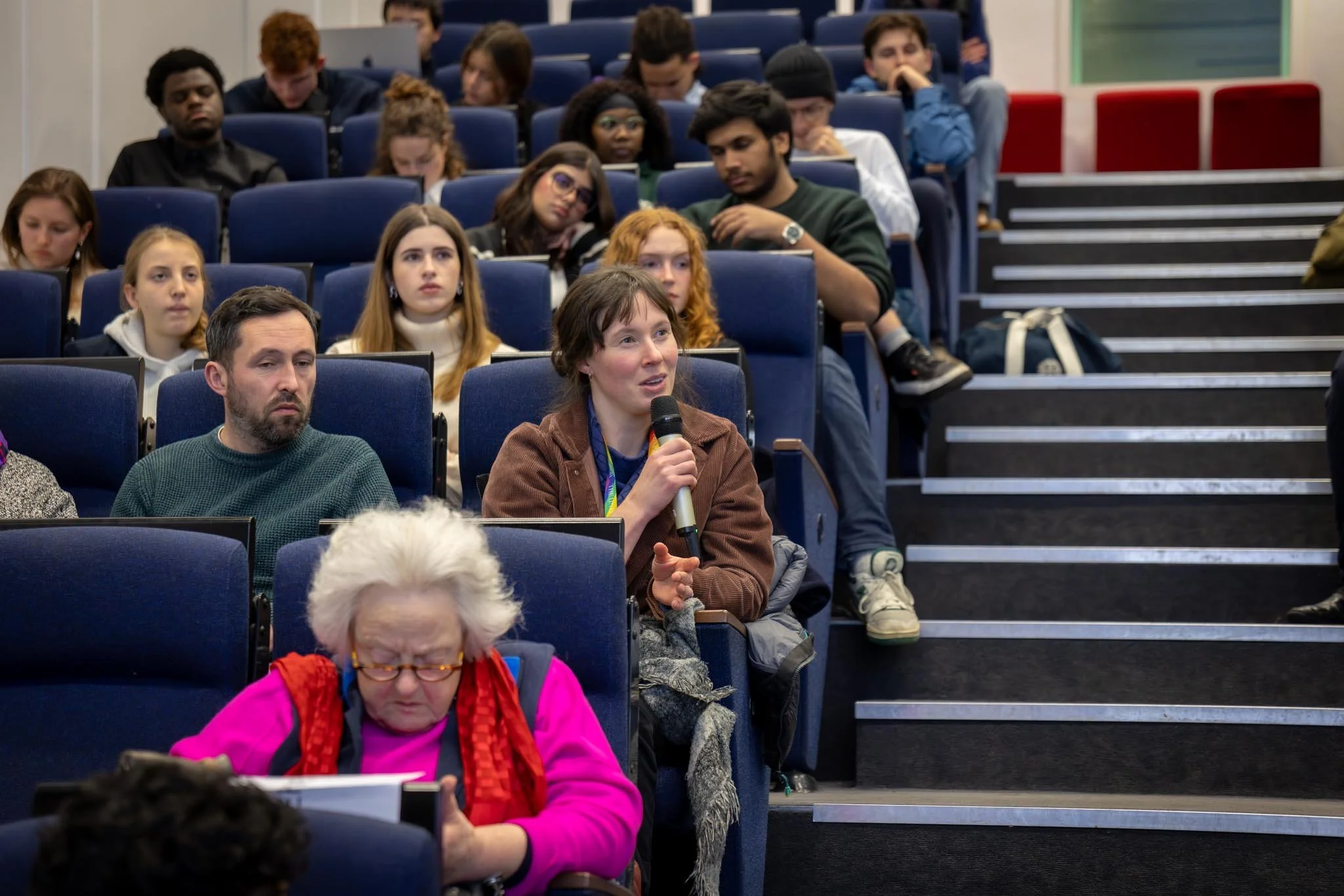 A woman holding a microphone speaking during a conference or seminar, seated in a blue theater-style auditorium with other attendees around her, including an elderly woman in the foreground and a diverse group of young adults in the background.