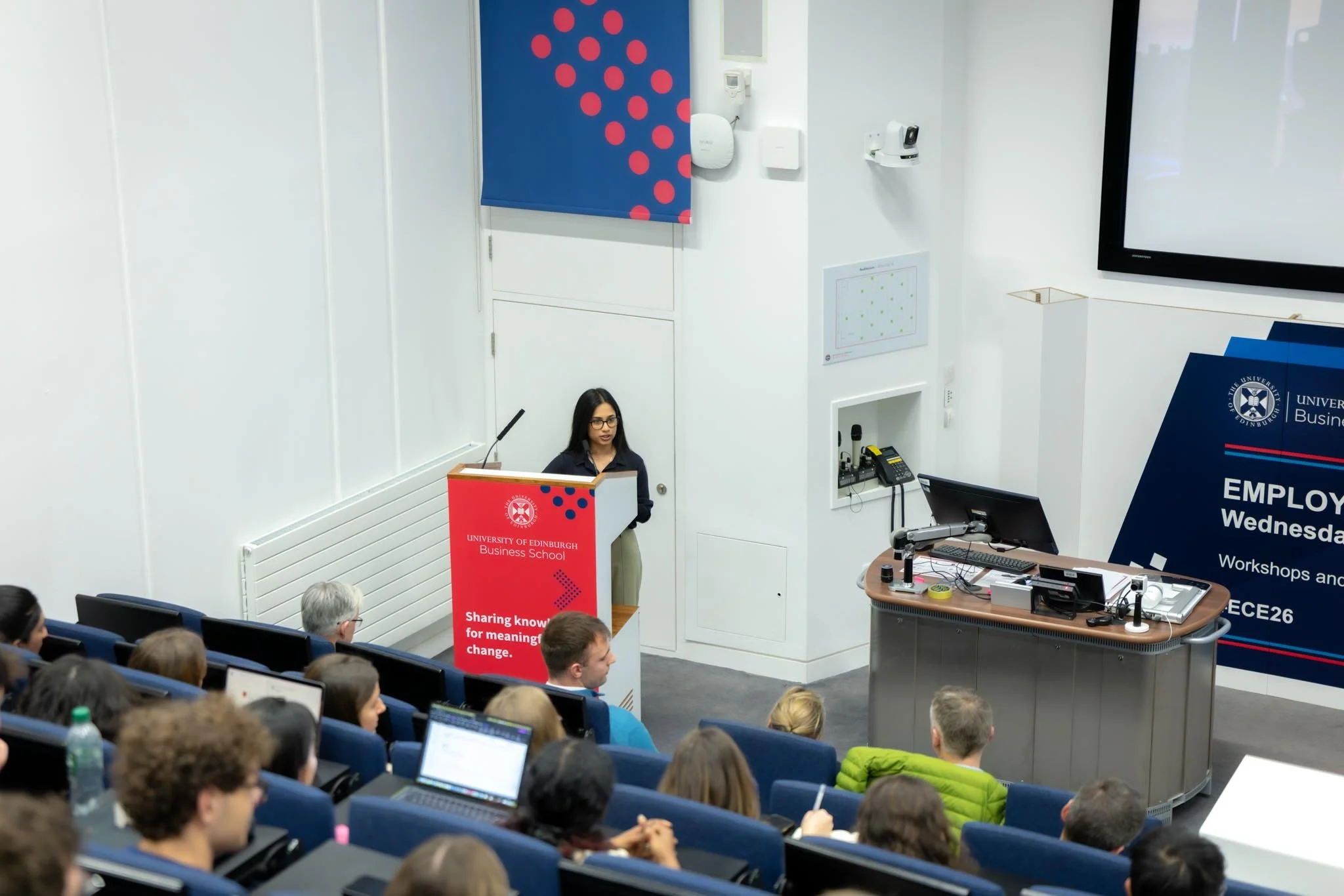 A woman giving a presentation at the University of Edinburgh Business School, with an audience seated in a lecture hall.