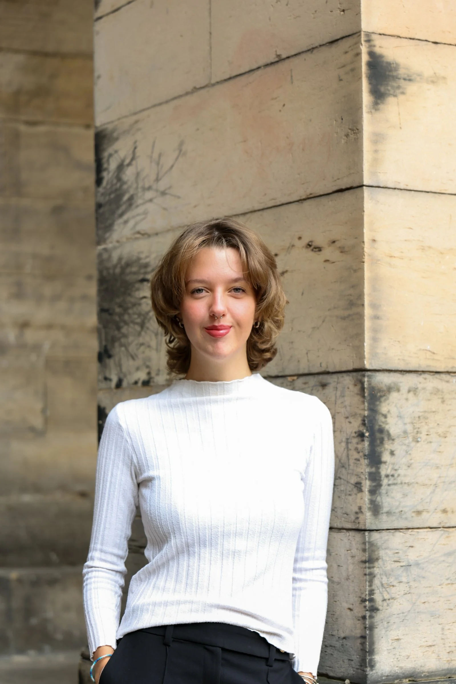 A young woman with short wavy hair wearing a white long-sleeve shirt and black pants standing against a stone building wall with a slight smile.