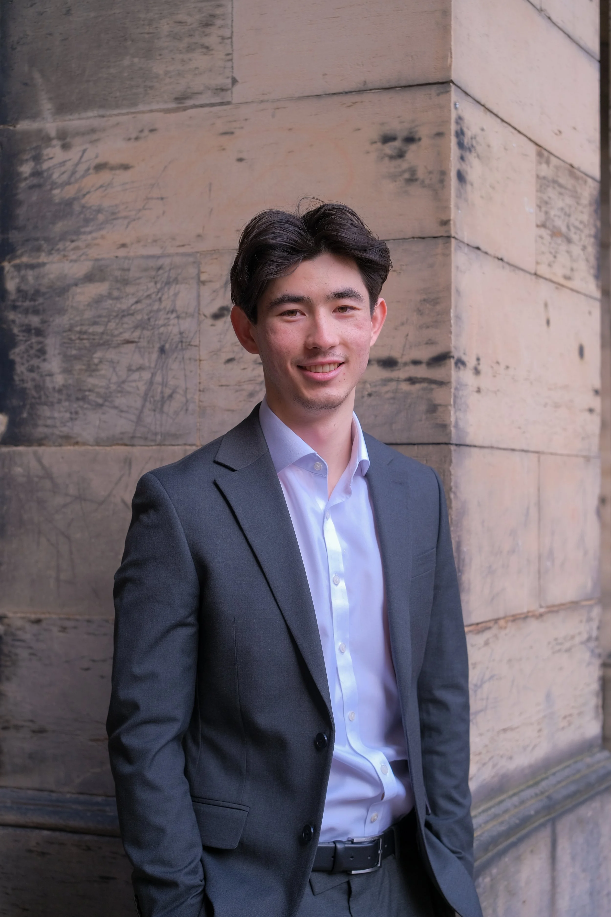 A young man in a gray suit and white shirt standing outdoors near a stone building.